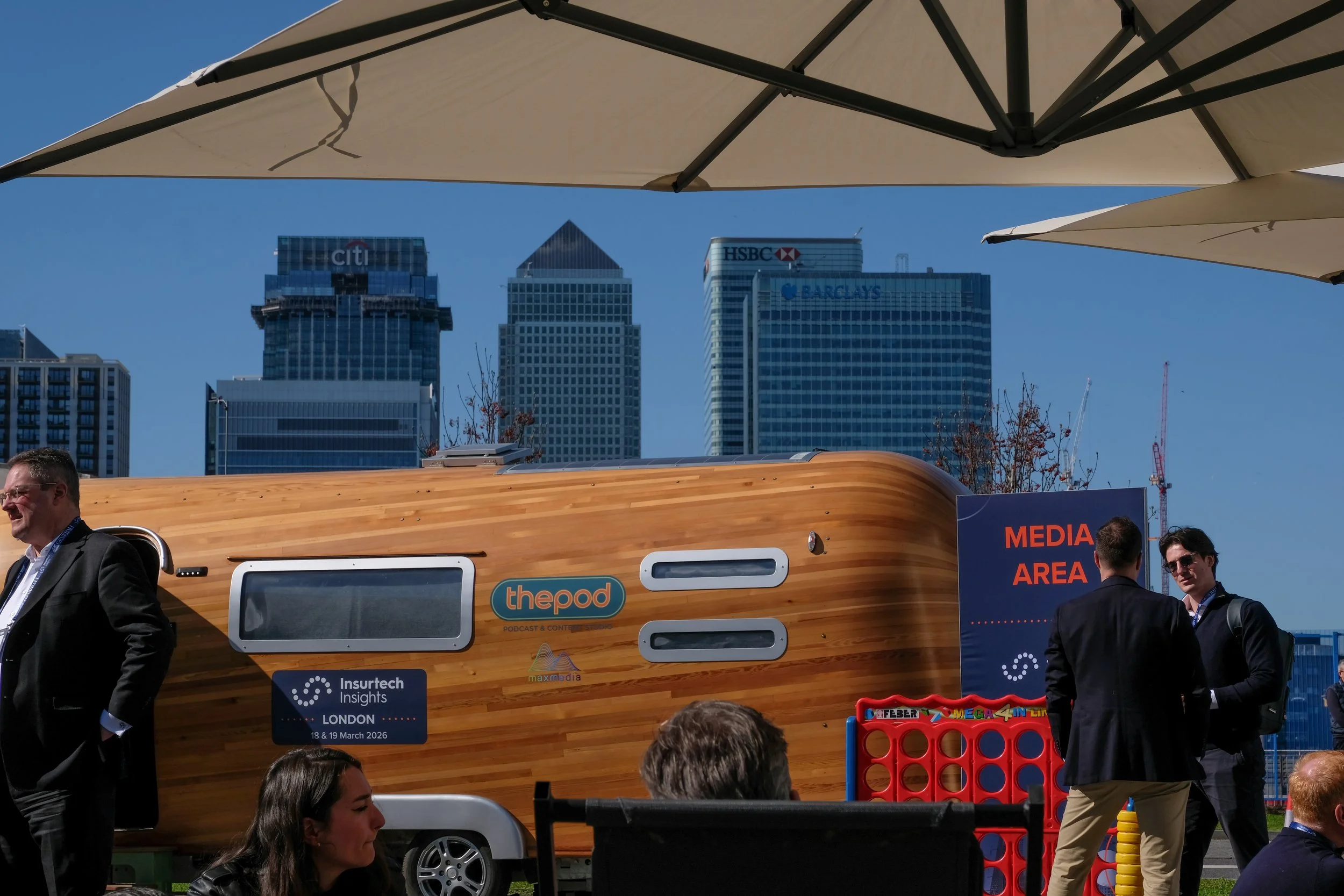 People gathered outdoors at a media area during a conference, with tall city buildings and a blue sky in the background. There is a wooden structure with various signs on it, including one for the podcast 'thepod' and event details for London on Marc