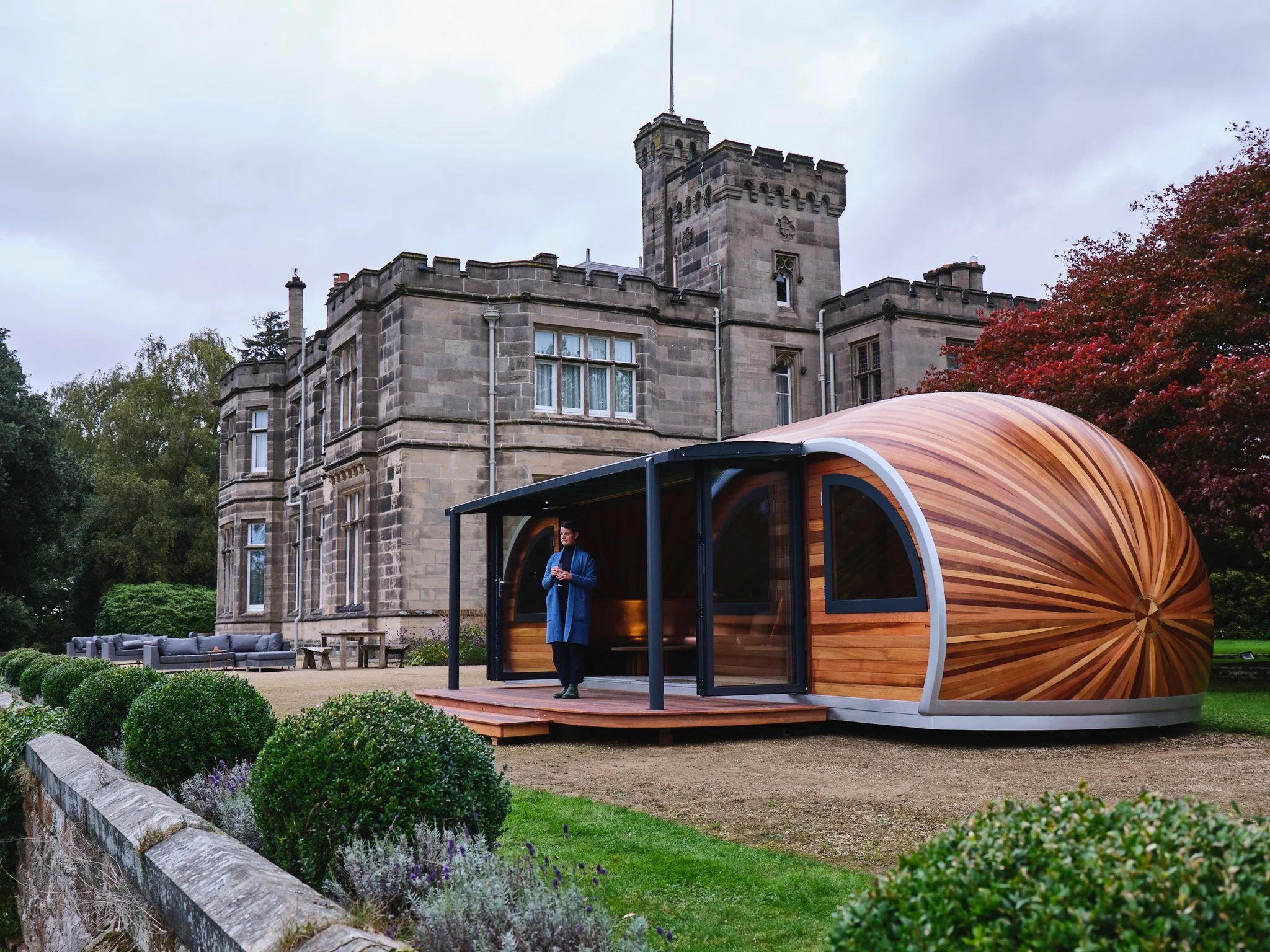 A woman standing inside a small, modern, curved wooden structure with a glass door, set on a lawn in front of an old stone castle with trees and outdoor furniture nearby.