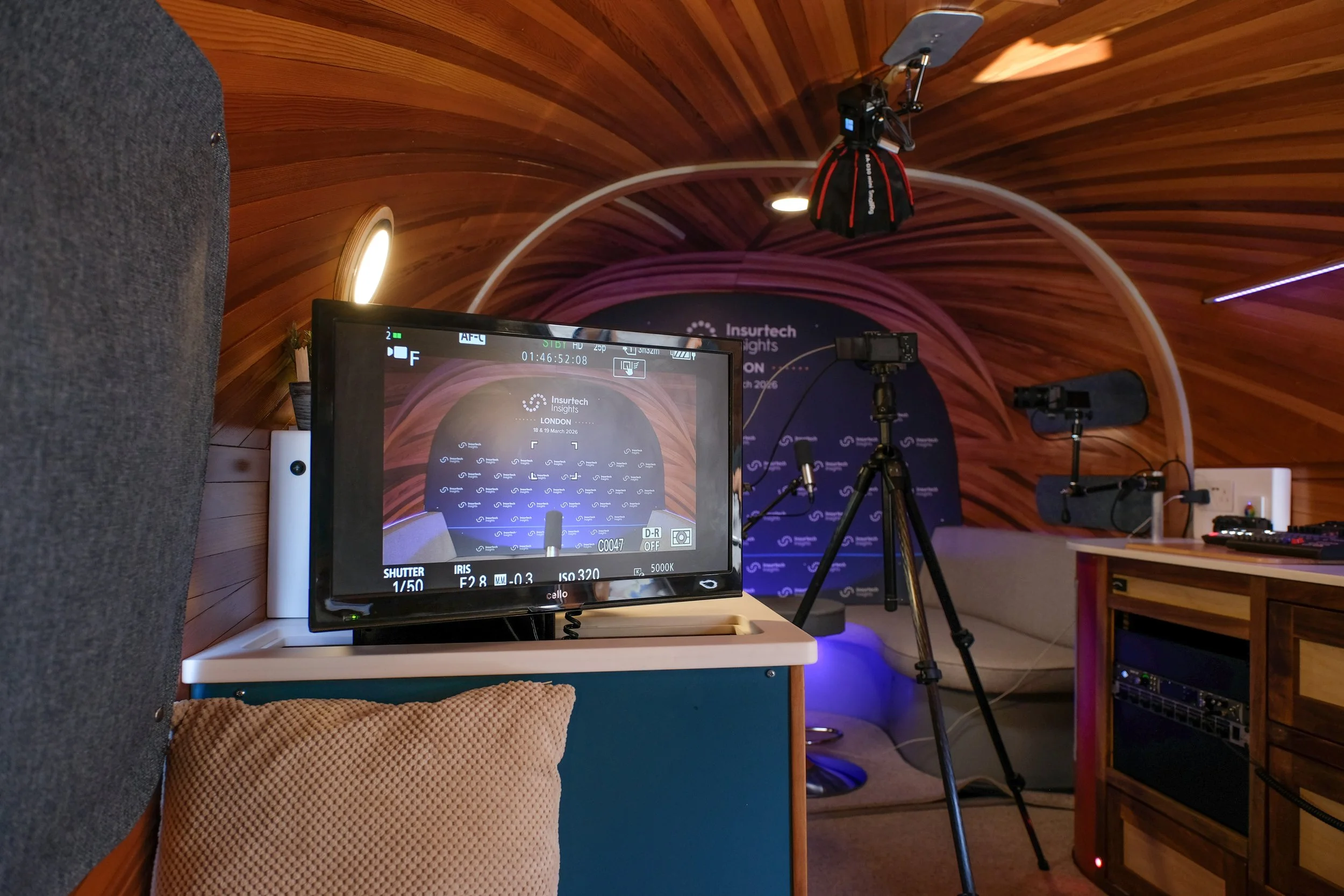 A camera monitor in the foreground shows a stage setup with a backdrop reading 'Insurtech Insights London 18 & 19 March 2026.' The background features a wooden arched ceiling, lights, tripods, and audio equipment in a conference or event room.