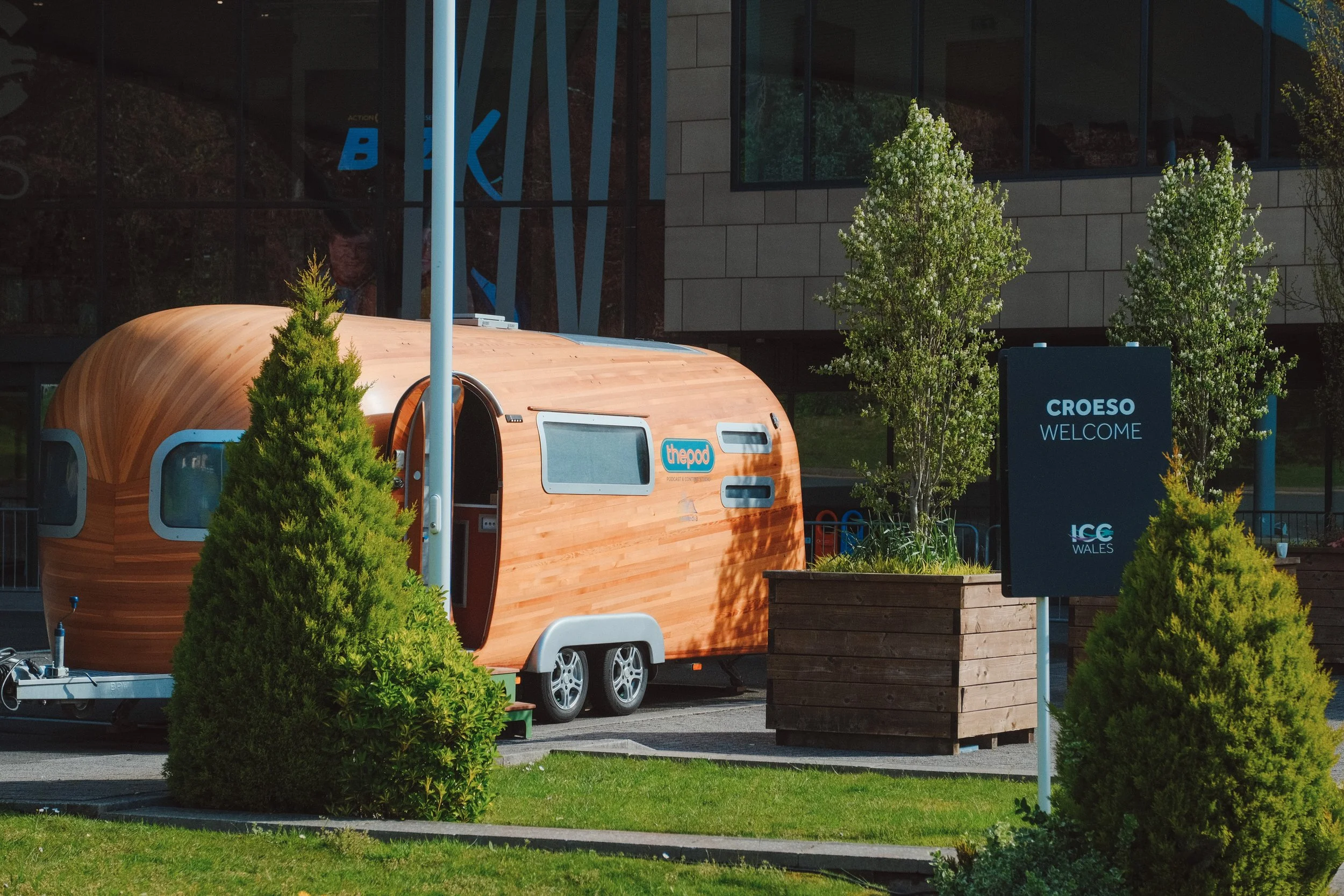 A small wooden trailer resembling a tiny house is parked in an outdoor area with green shrubs and trees. There is a sign that says 'CROESO WELCOME' and some other text on it, and a larger building with glass windows reflects some trees.
