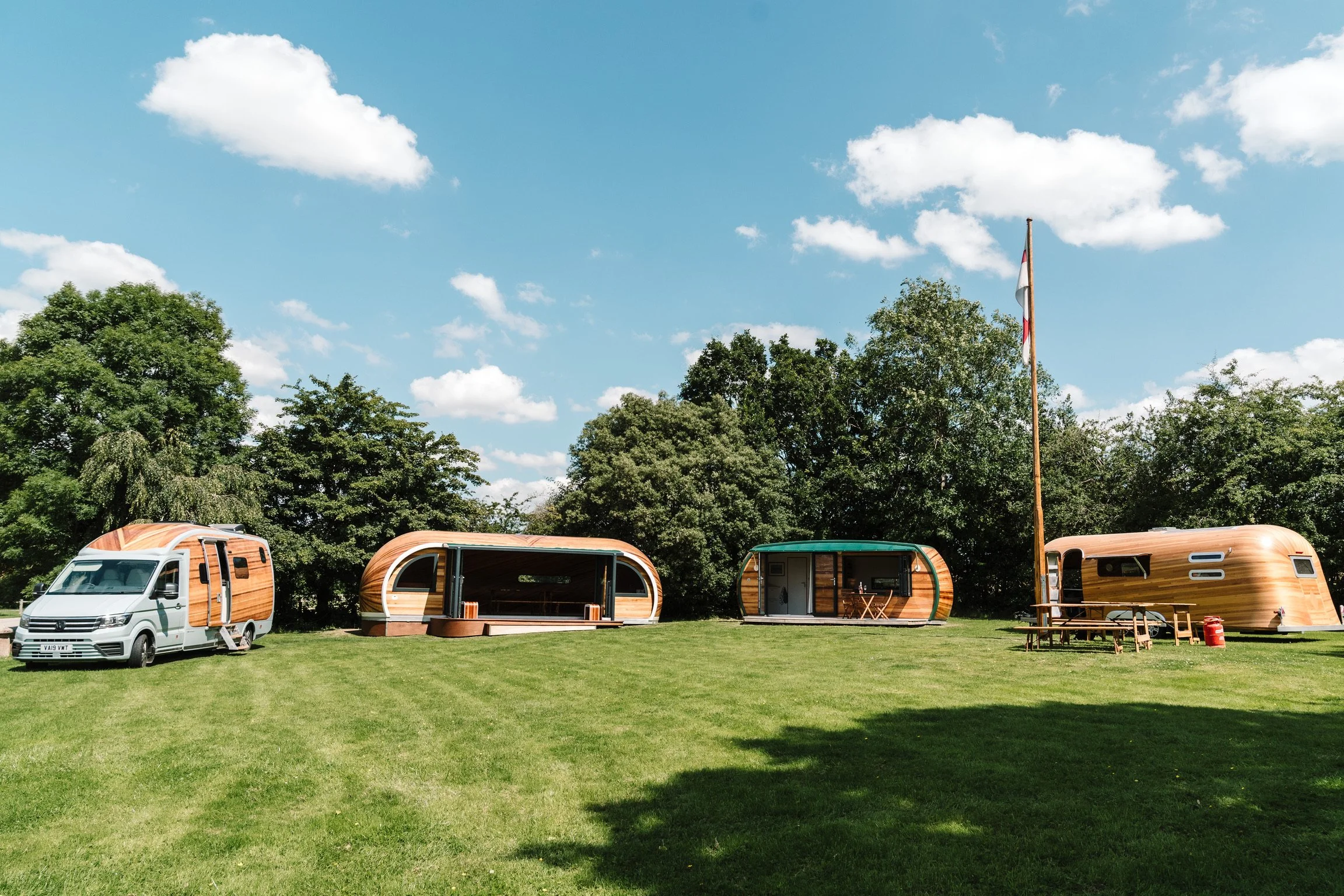 A grassy field with four wooden camping trailers, one with a small outdoor dining set, set against a backdrop of tall trees and a partly cloudy sky, with a flagpole flying a white and red flag.