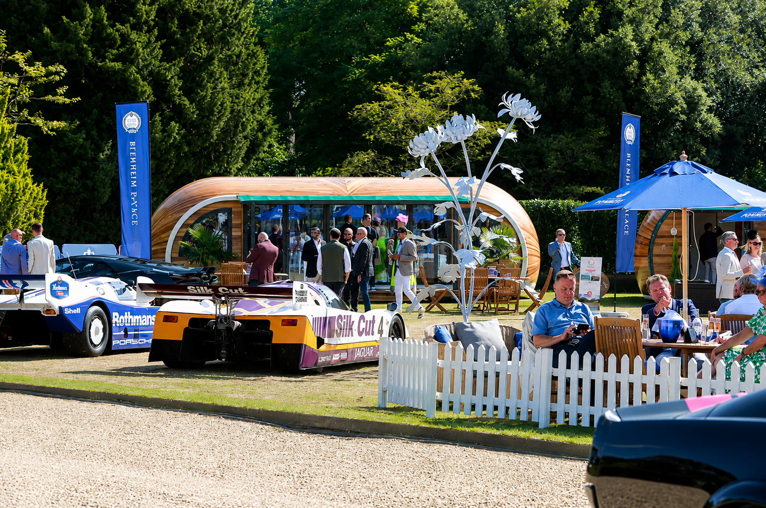 People socializing outdoors at a car exhibition with vintage race cars, modern structures, blue umbrellas, and lush green trees in the background.
