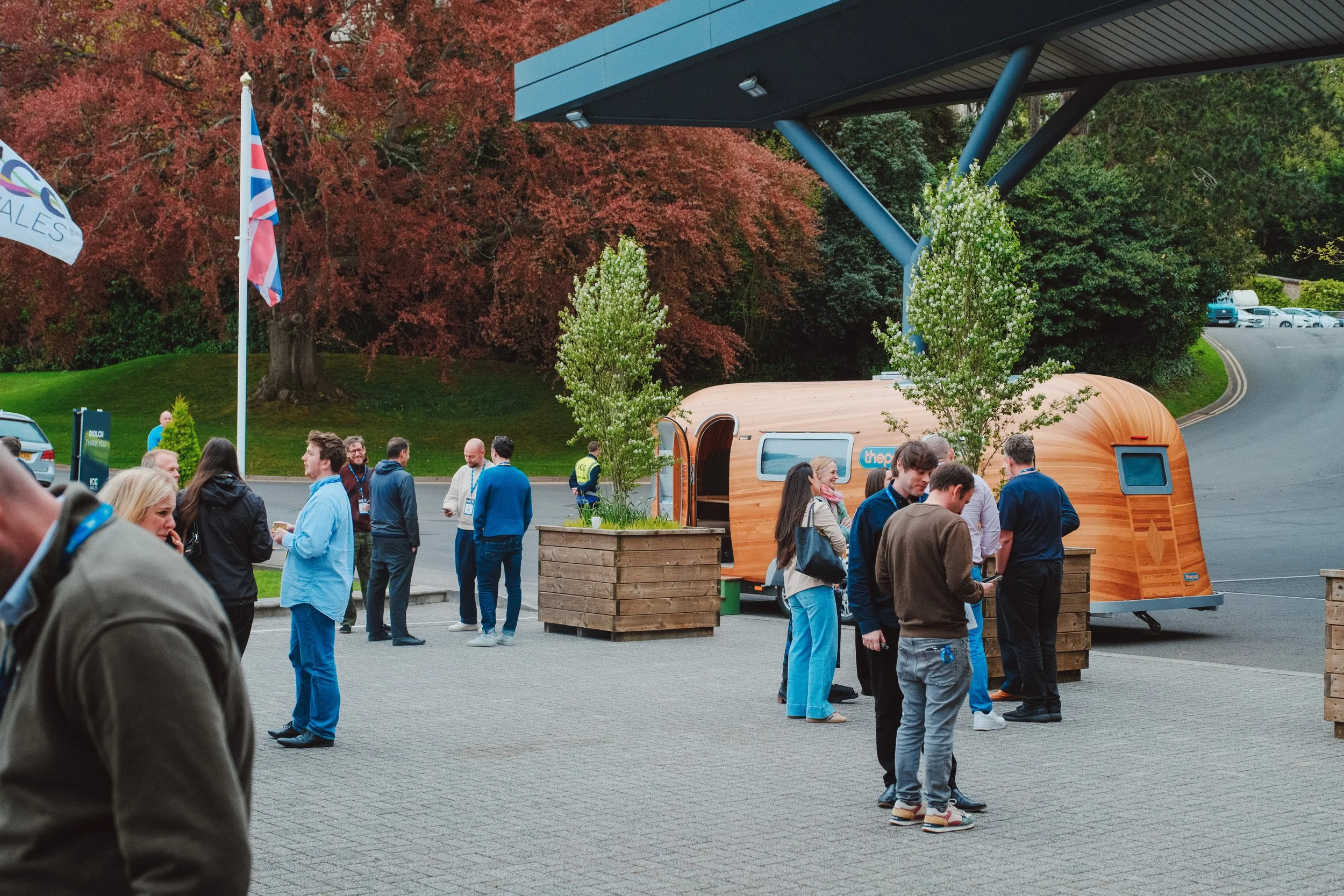 People gathered outdoors near a small wooden food truck, with trees and parking lot in the background, during daytime.