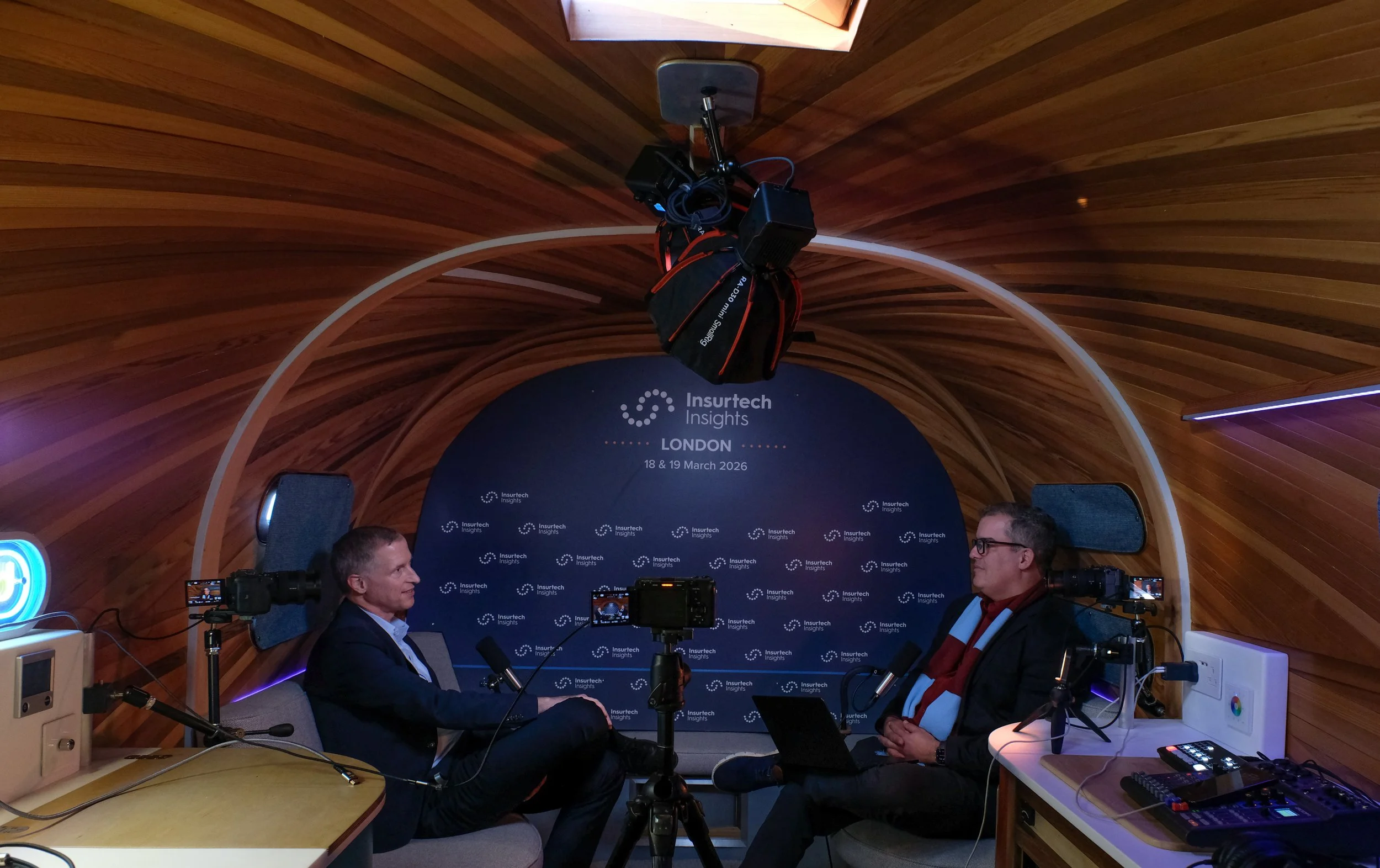 Two men sitting in a wooden, curved room with a blue backdrop that reads 'Insurtech Insights London 18 & 19 March 2026.' They are engaged in a discussion, surrounded by cameras, microphones, and other equipment, with a camera and a microphone on a de