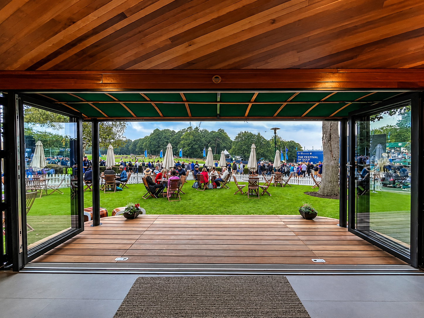 Open patio door looking out onto a golf event with people seated on green grass and umbrellas, a white picket fence, trees, and a sign that reads 'The Festival of Golf'