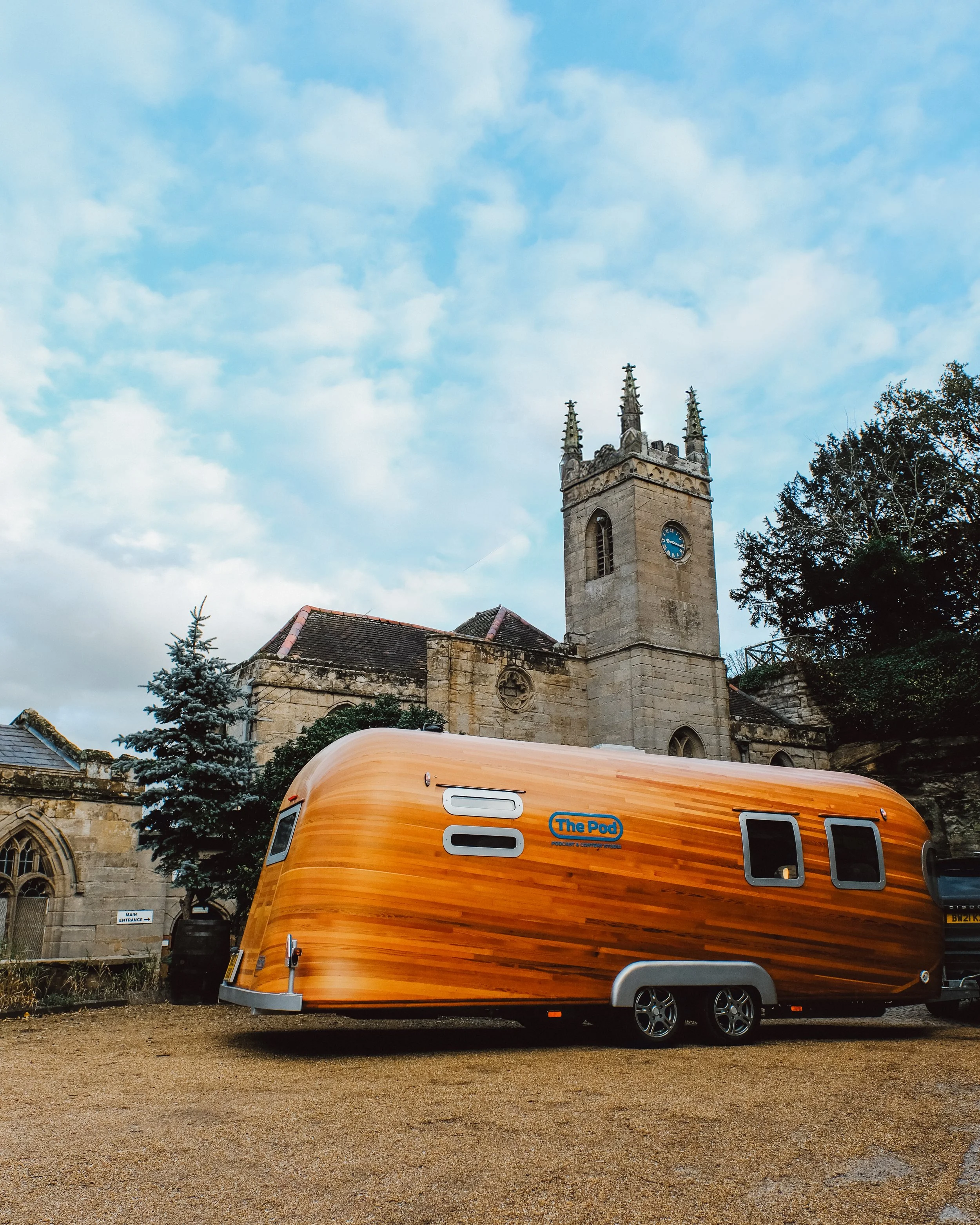 A modern wooden RV trailer parked in front of a historic church with stone walls, clock tower, and gothic windows, with trees and cloudy sky in the background.