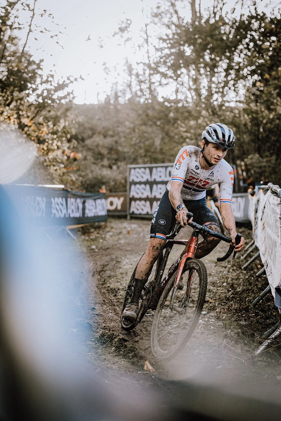 A cyclist wearing a white helmet and a white jersey with orange and blue accents, riding a black and orange bike through a muddy, wooded off-road course during a race, with barriers and banners in the background.