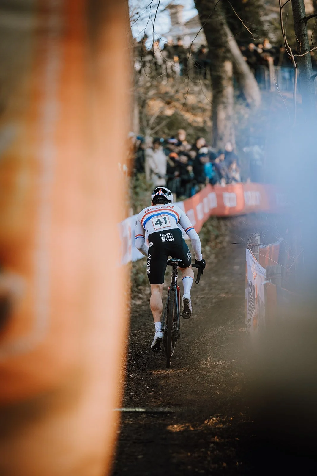 Cyclist riding downhill on a forest trail during a race, viewed from behind with spectators in the background.