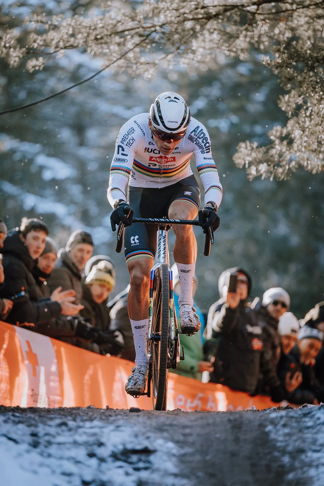 A cyclist competing in a race on a muddy trail, with spectators watching along the side.