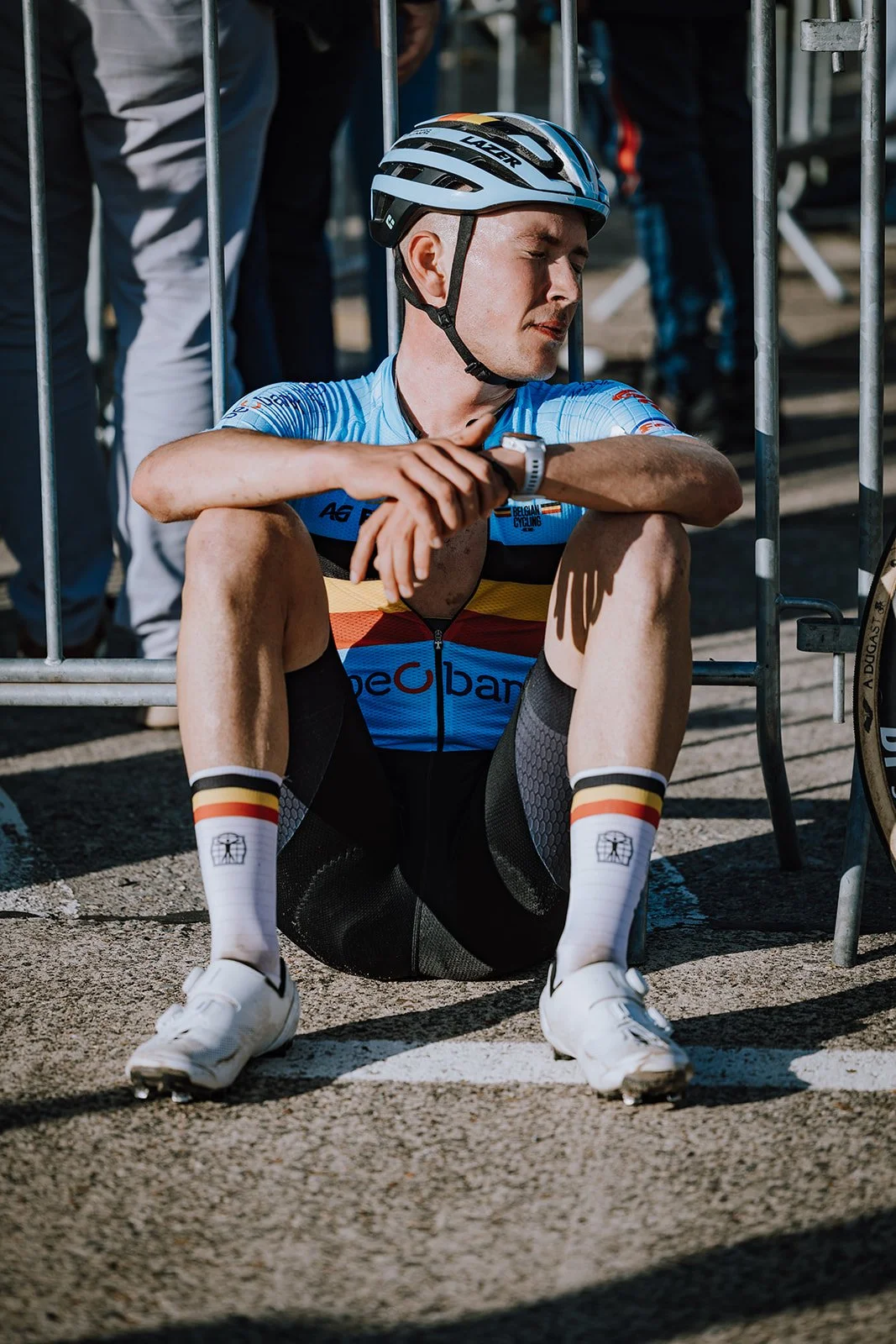 A male cyclist sitting on the ground, wearing a cycling helmet, jersey, shorts, and socks, looking at his watch with a concerned expression after a race.