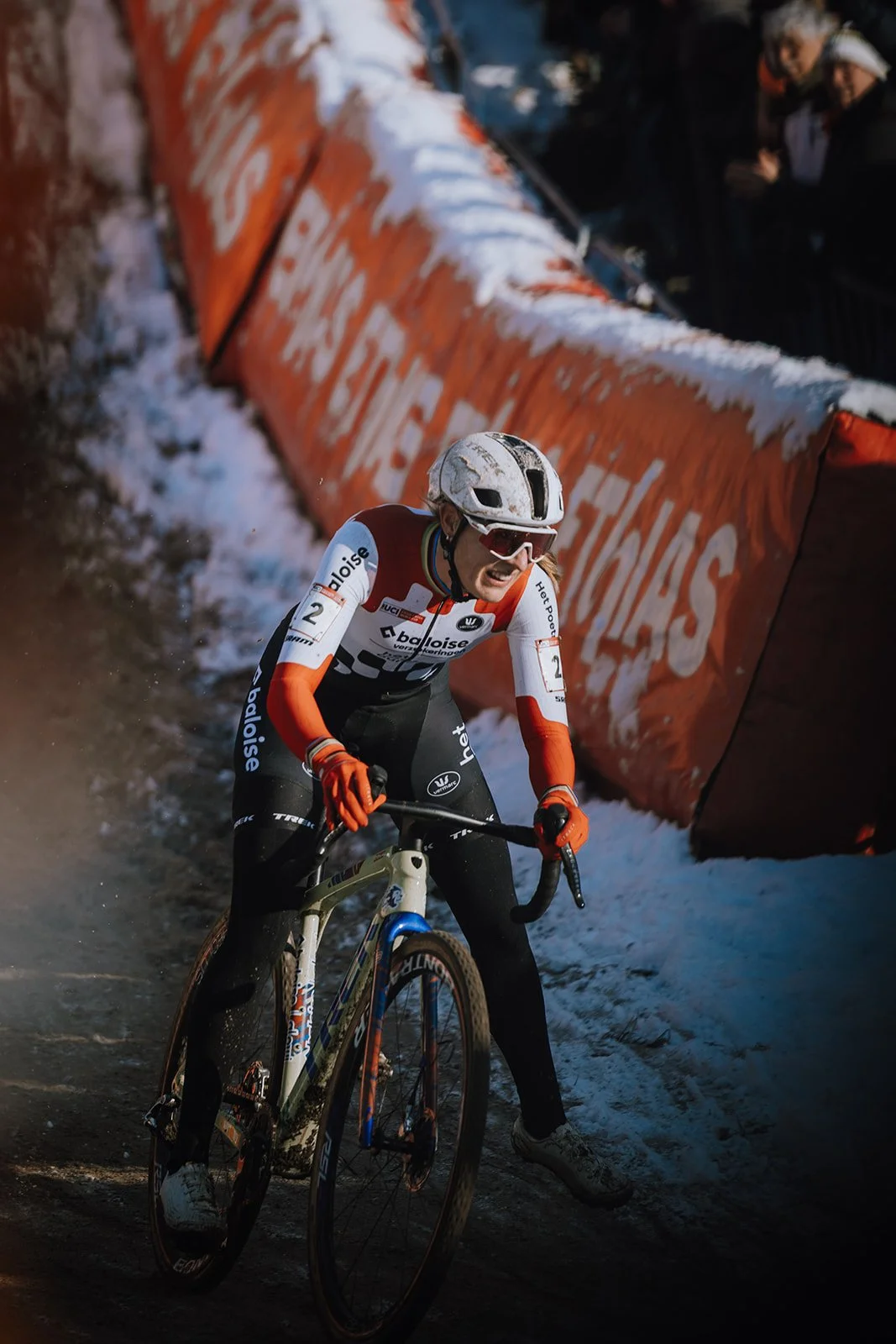 A female cyclist wearing a helmet, goggles, orange gloves, and a team jersey, racing on a muddy and snowy outdoor track with an orange barrier in the background.