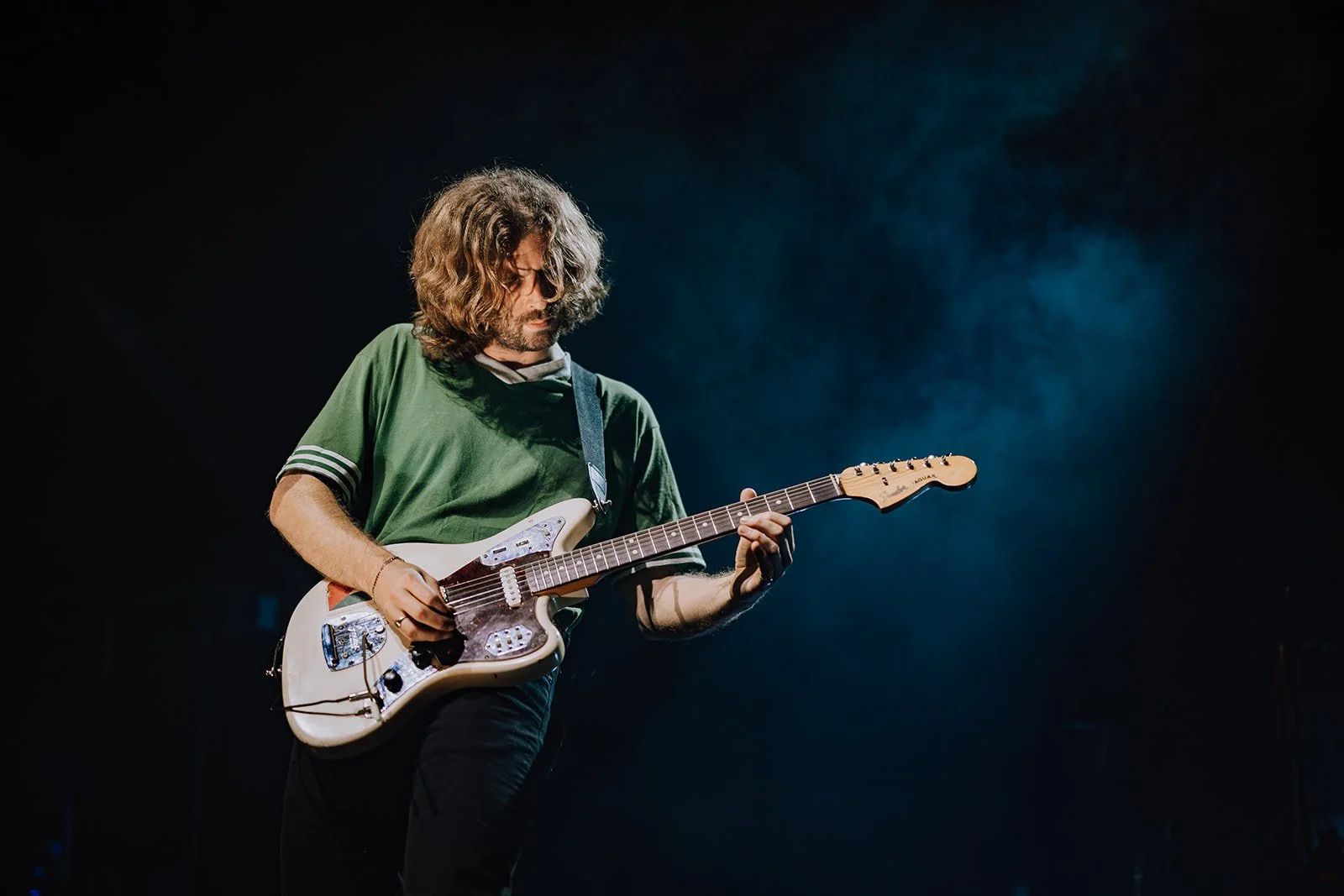 A man with shoulder-length curly hair playing an electric guitar on stage with a dark background and subtle lighting.