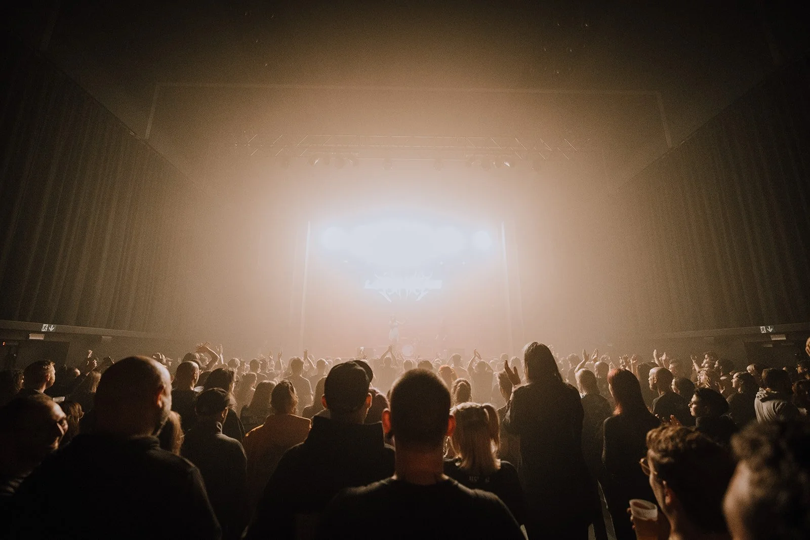 Crowd of people at a concert or music event facing a brightly lit stage with an artist performing.