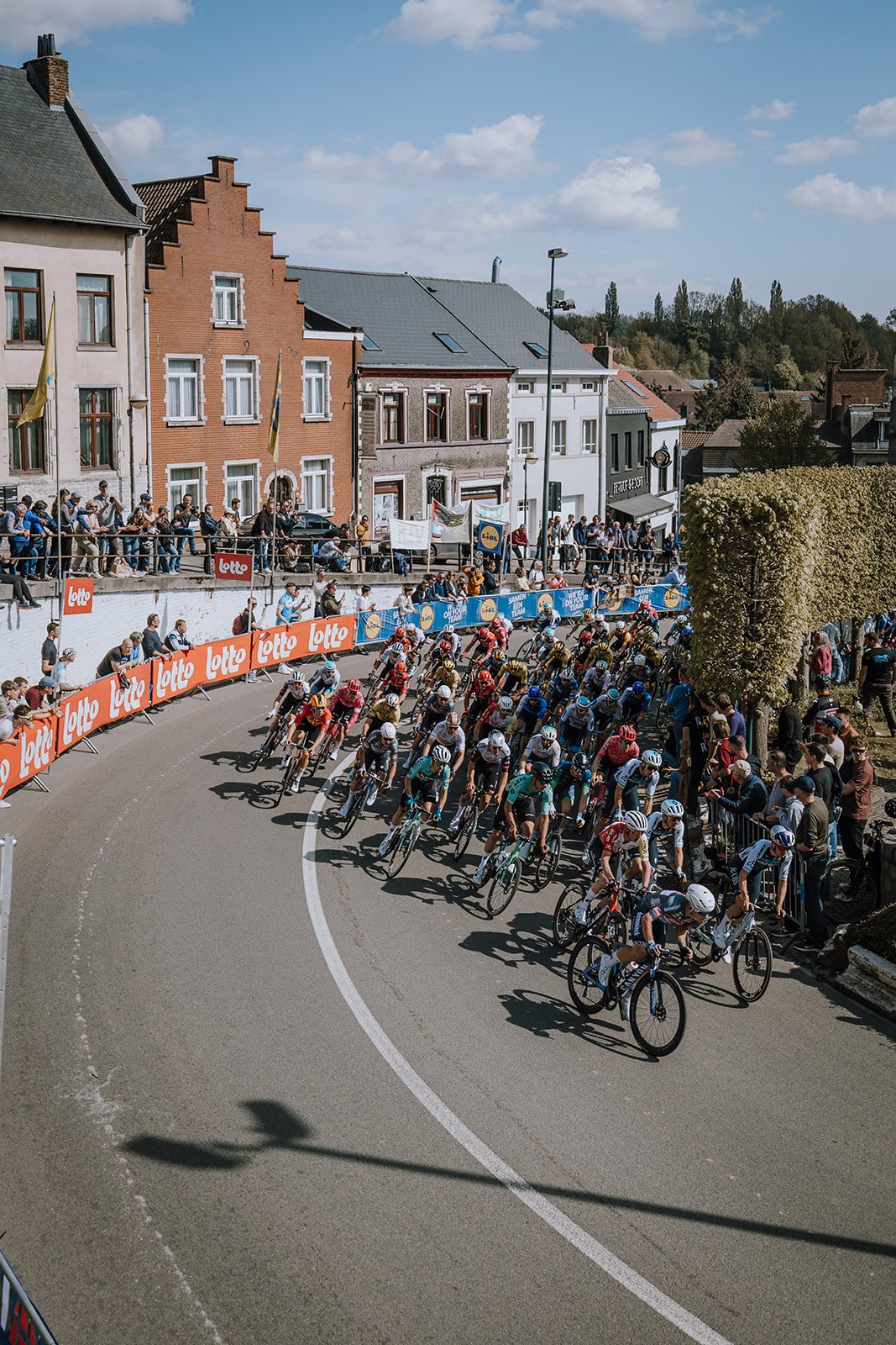 A cycling race taking place on a city street with a large group of cyclists, spectators along the sidewalk, and banners advertising Lotto and other sponsors.