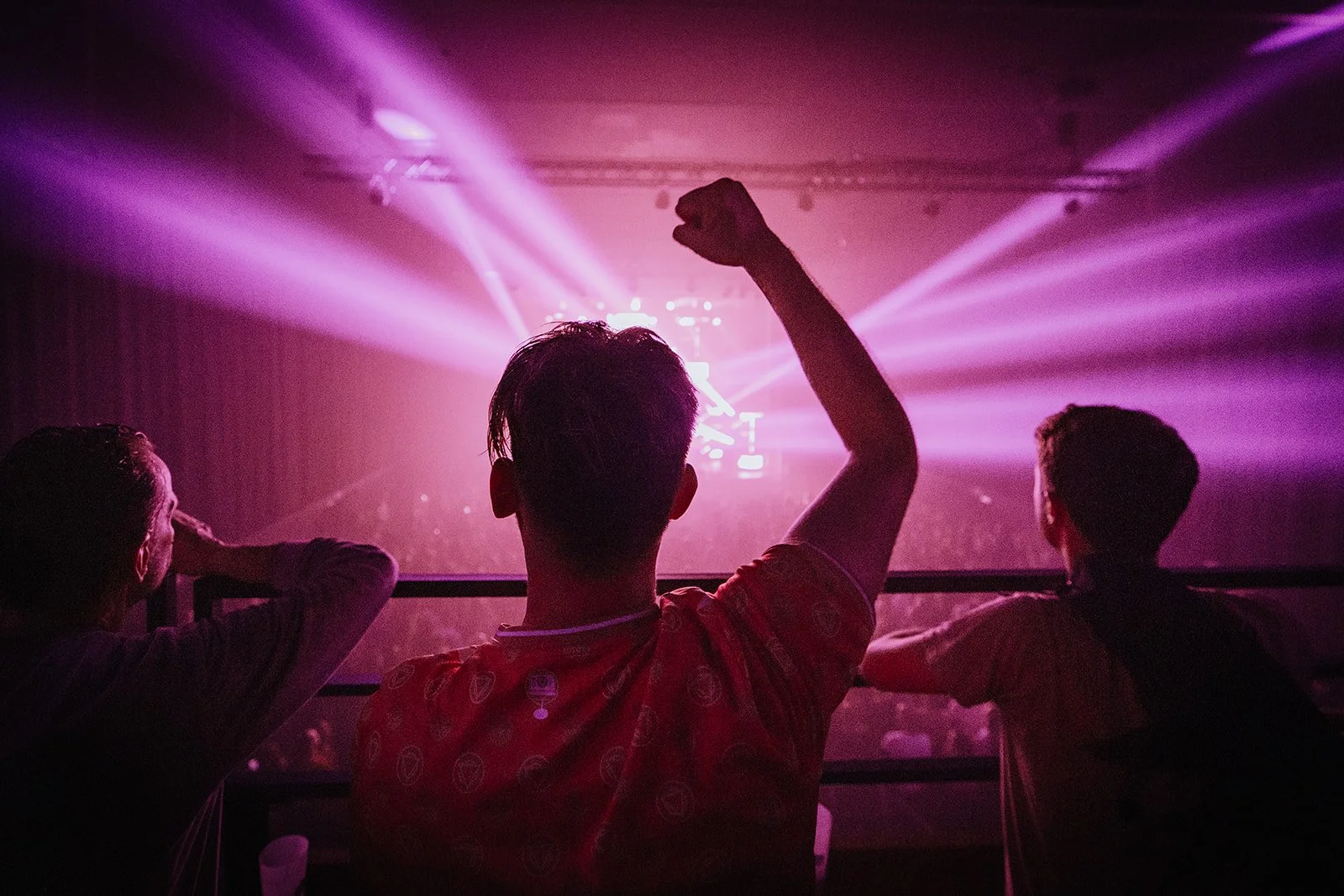 People sitting in an audience at a concert or event with purple and pink lights illuminating the stage, one person raising a fist in excitement.