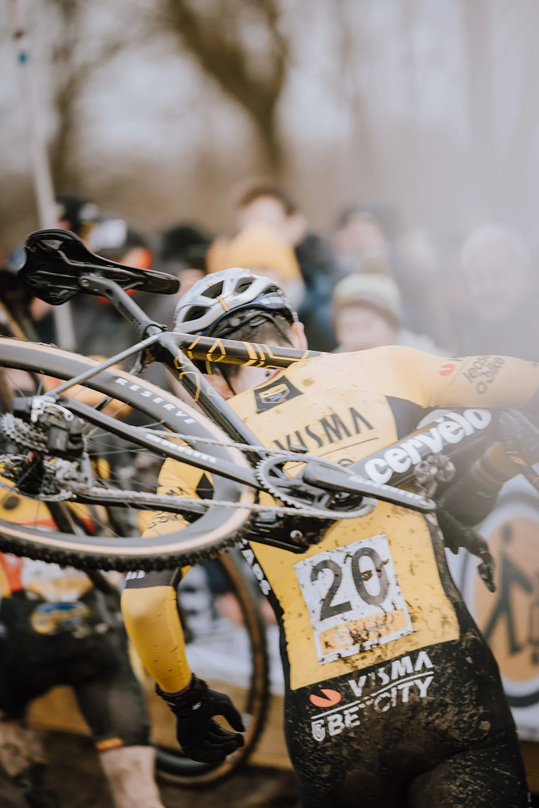 Mountain biker in yellow jersey carrying bike on his shoulder, muddy race setting, crowd of spectators in the background.