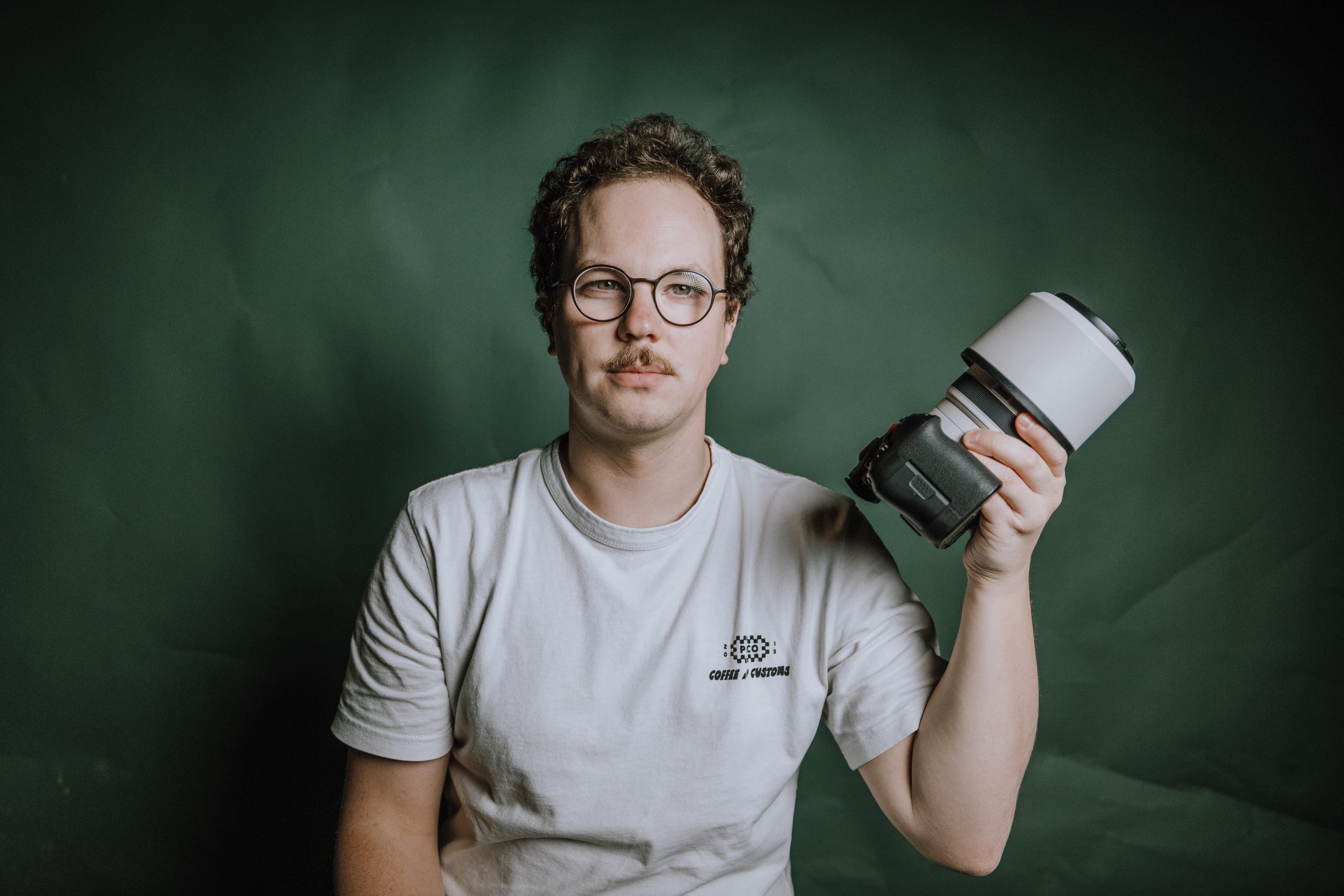 A person with glasses, mustache, and curly hair holding a professional camera with a large lens, standing against a green background.