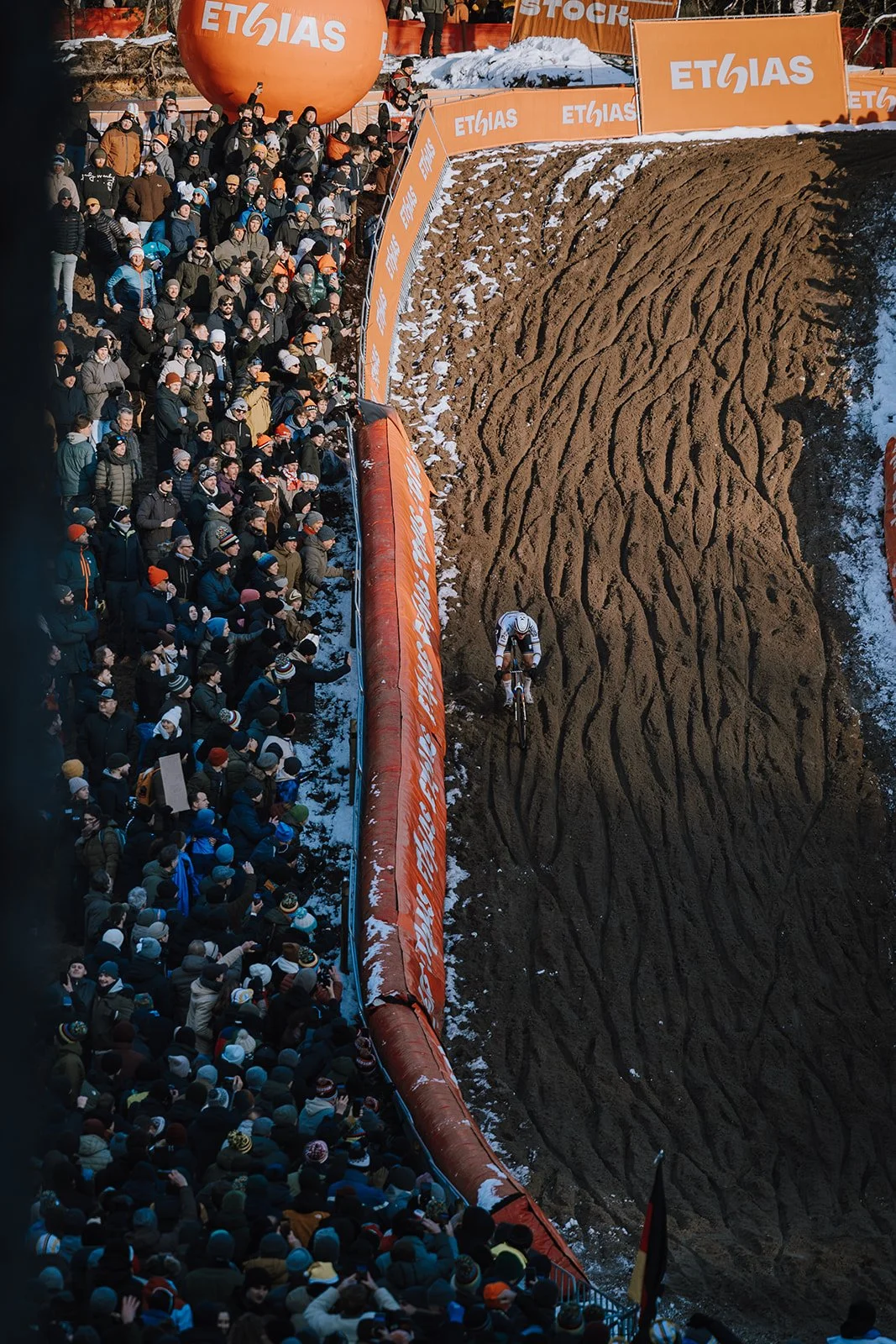 A cyclist riding on a dirt track during a race, with a large crowd of spectators watching from the side, festival banners, and snow on the ground.
