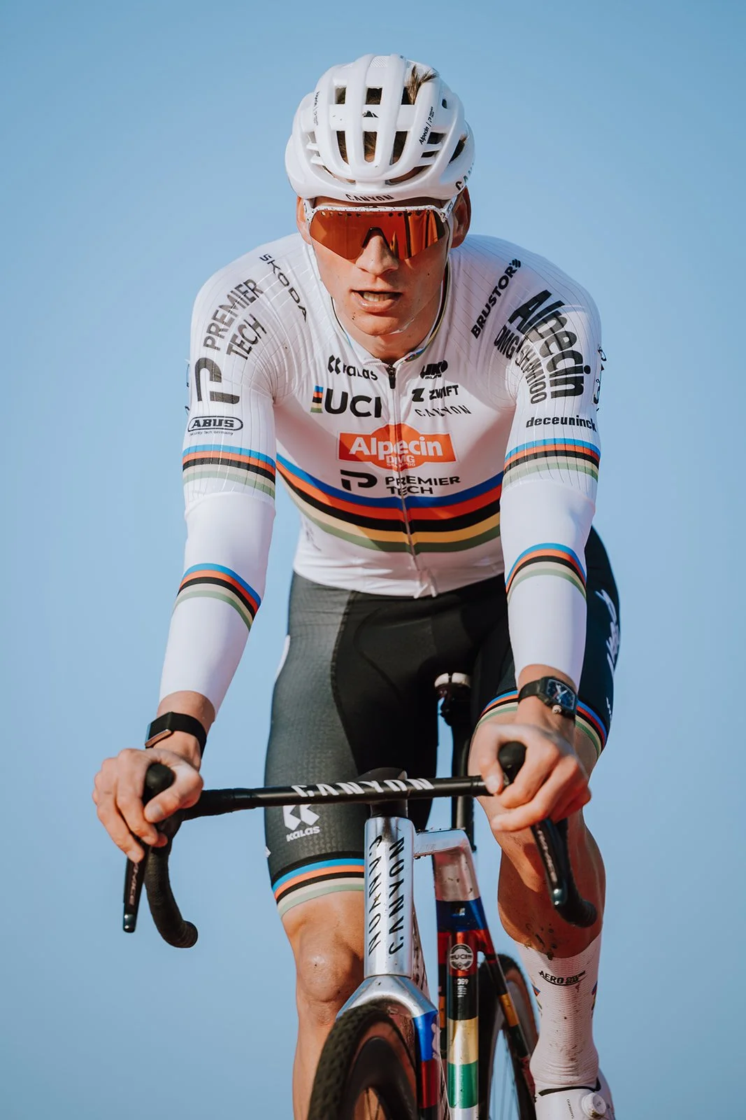 Male cyclist wearing a white racing jersey, black shorts, and a white helmet, riding a bike against a clear sky.