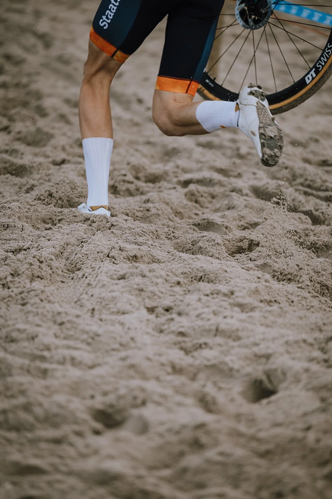 Close-up of a cyclist's legs and feet as they ride through sand on a beach, wearing white socks and athletic shoes, with part of a black and orange cycling uniform and a bicycle visible.