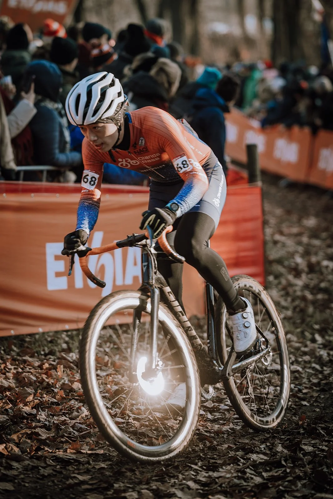 A cyclist wearing a white helmet, orange and black jersey with the number 68, leaning forward on a mountain bike during a race on a leaf-covered trail. Spectators line the course behind an orange barrier.