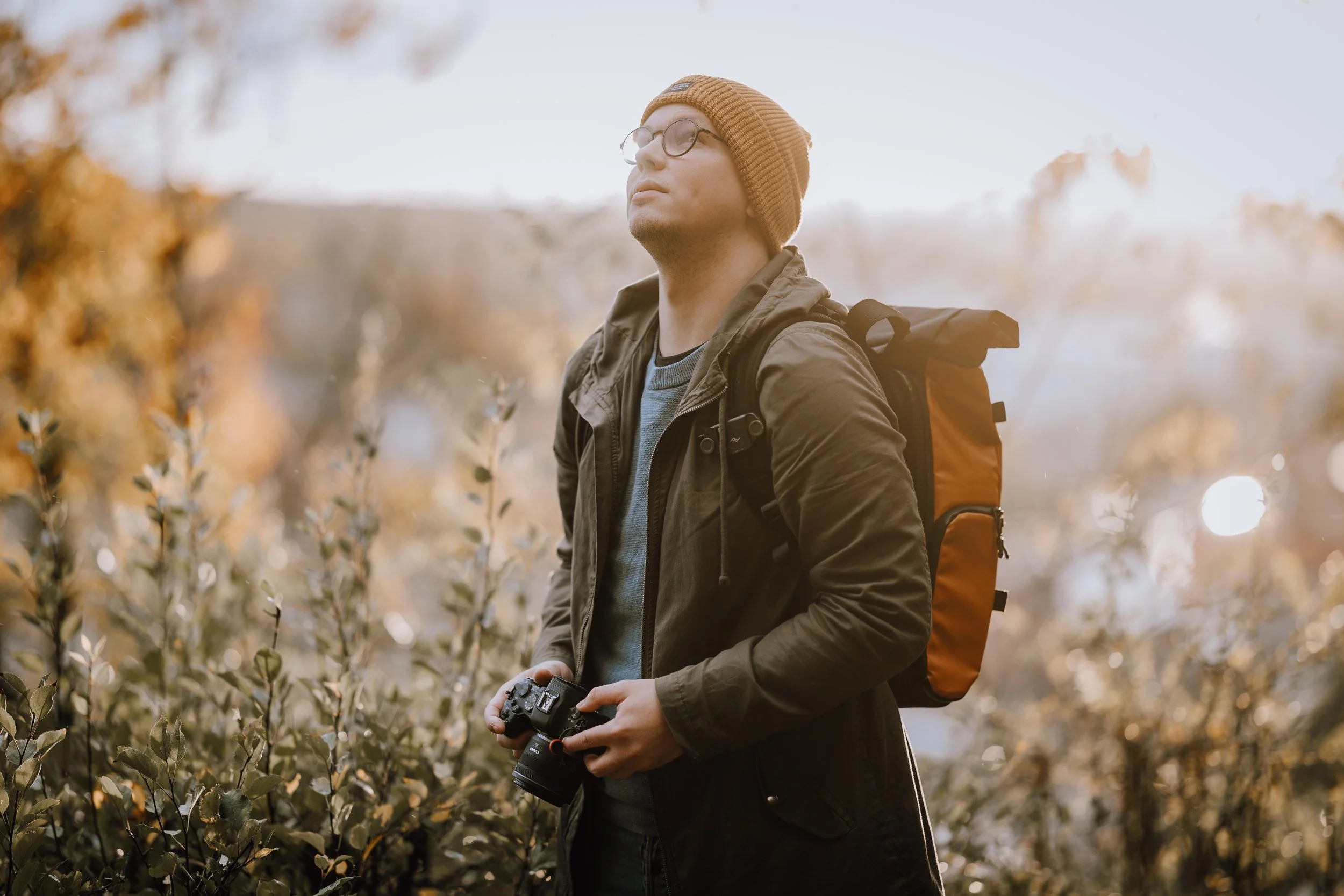 A man standing outdoors in autumn with a camera in his hands, wearing glasses, a brown beanie, a green jacket, and a backpack.