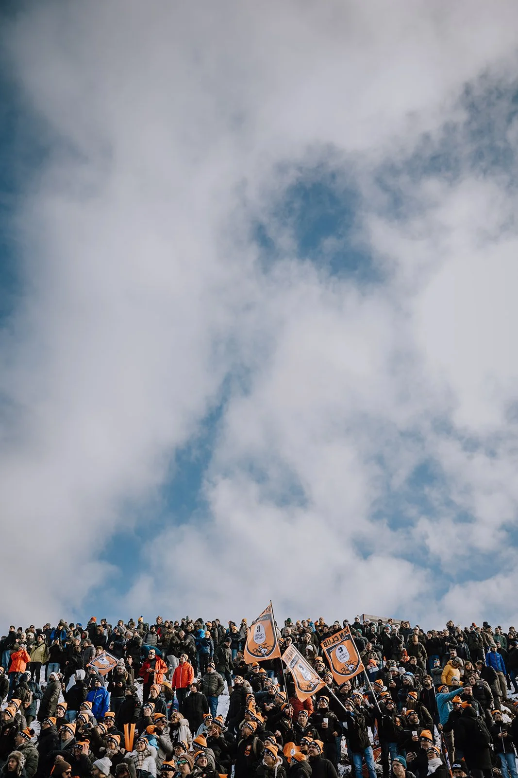 Crowd of people gathered outdoors, holding orange and black flags and banners, under partly cloudy sky.