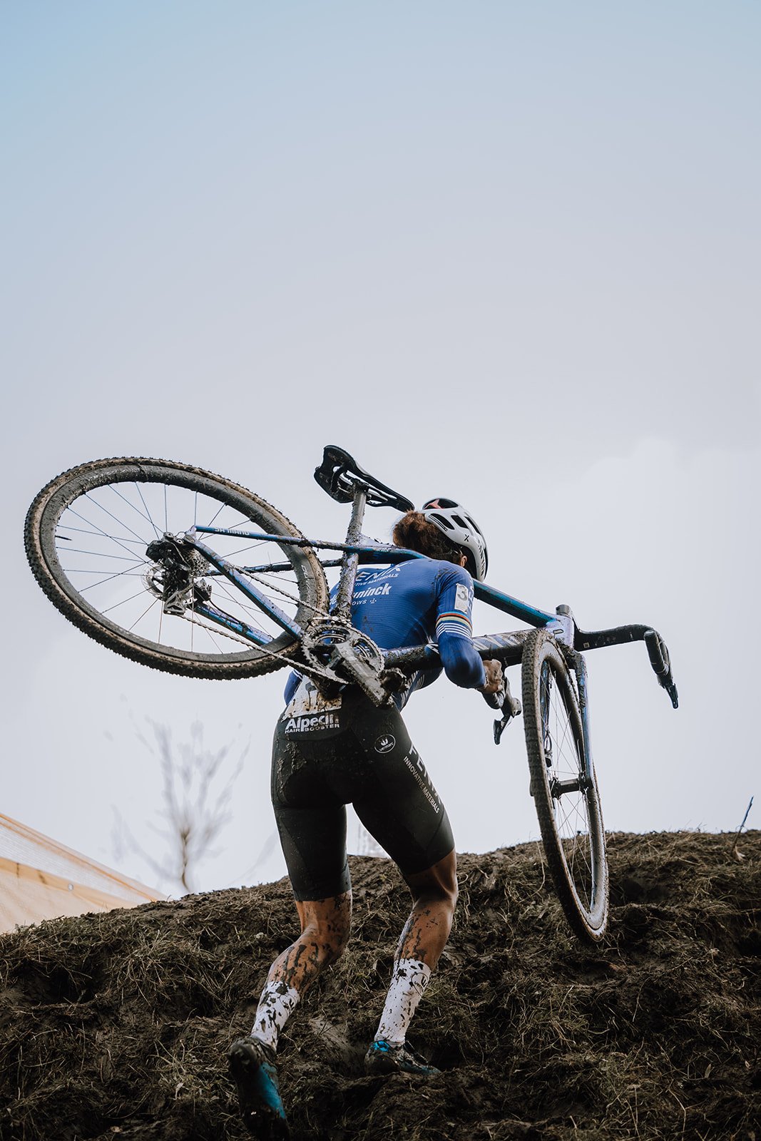 Cyclist carrying a muddy mountain bike uphill on a dirt trail during a cyclocross race.
