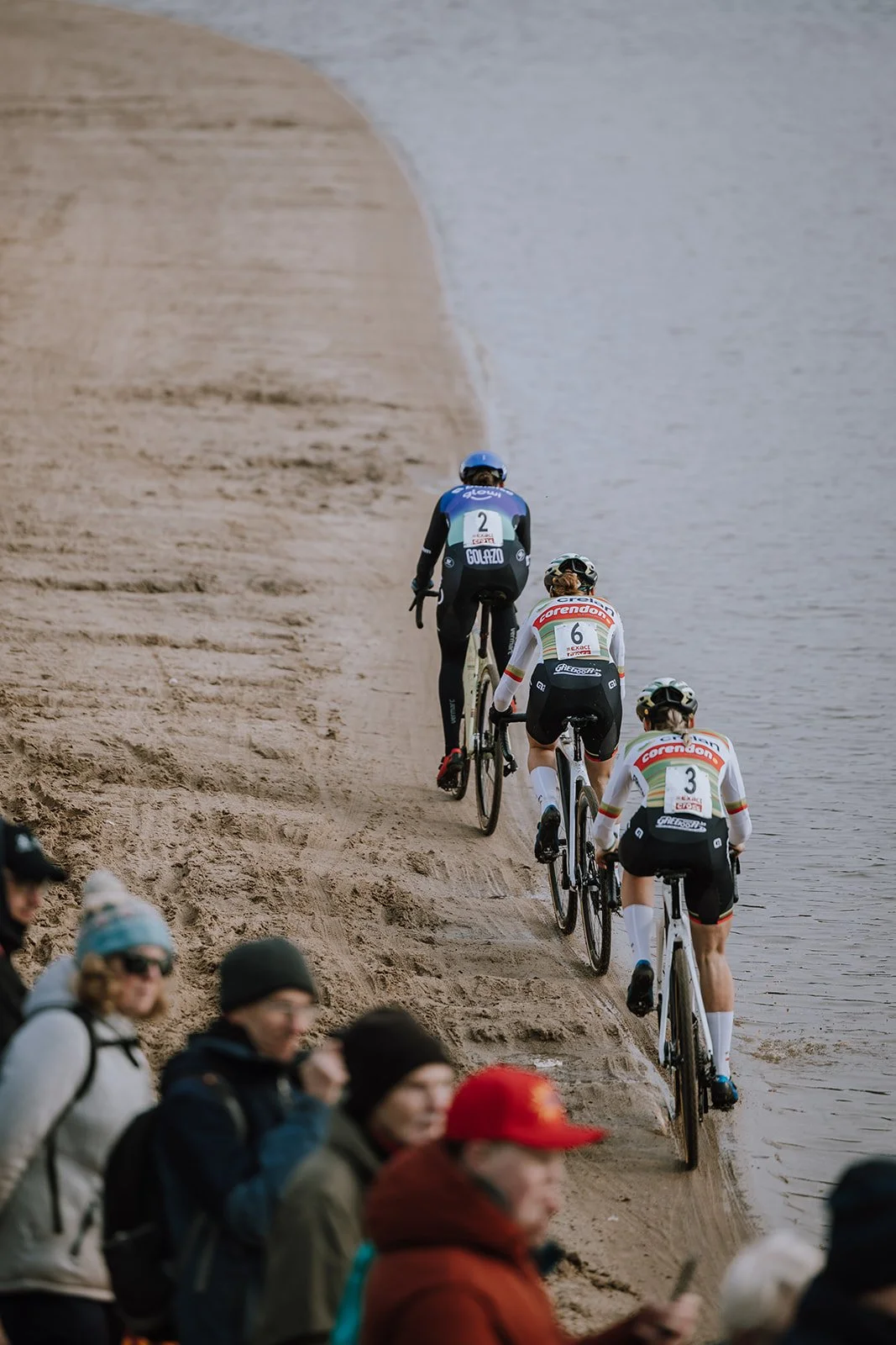 Cyclists riding along a sandy beach with spectators watching from the foreground