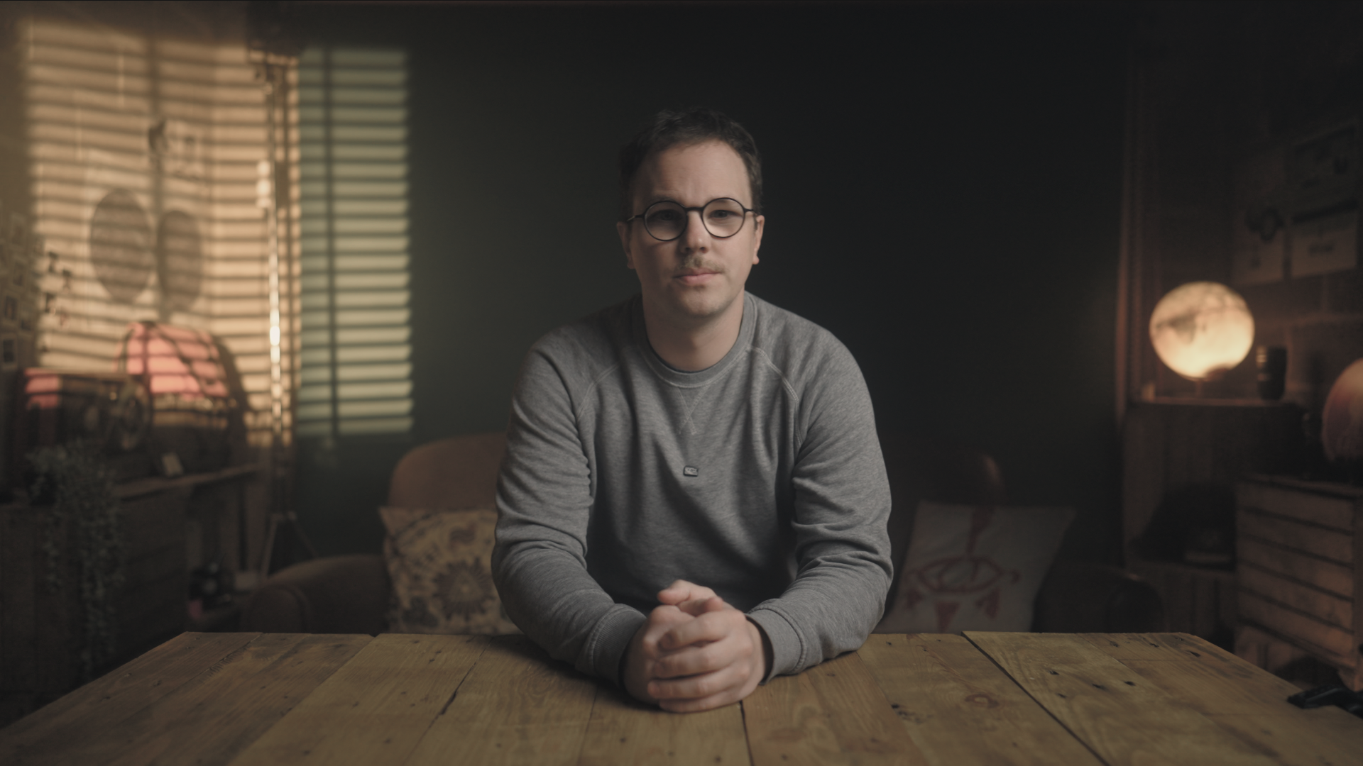 A man with glasses and a gray sweatshirt sitting at a wooden table in a dimly lit room with decorative lighting and blackboard behind him.