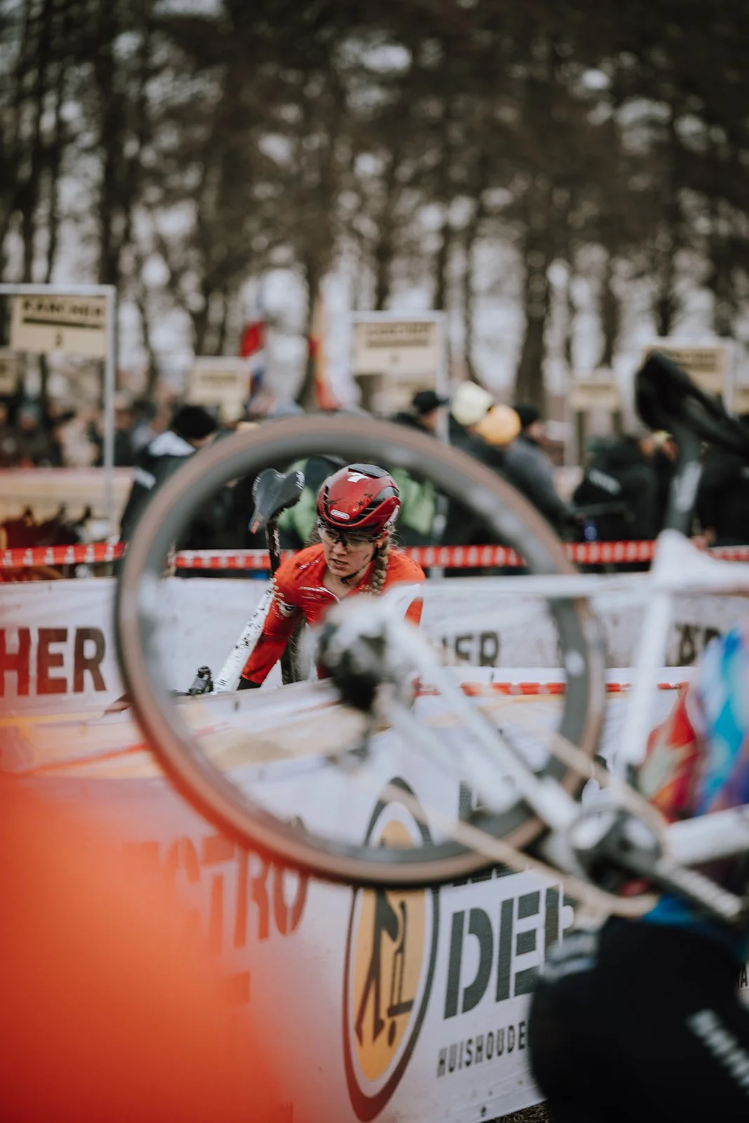 A cyclist wearing a red helmet and orange jersey at a cycling race, seen through a bicycle wheel in the foreground, with spectators and banners in the background.