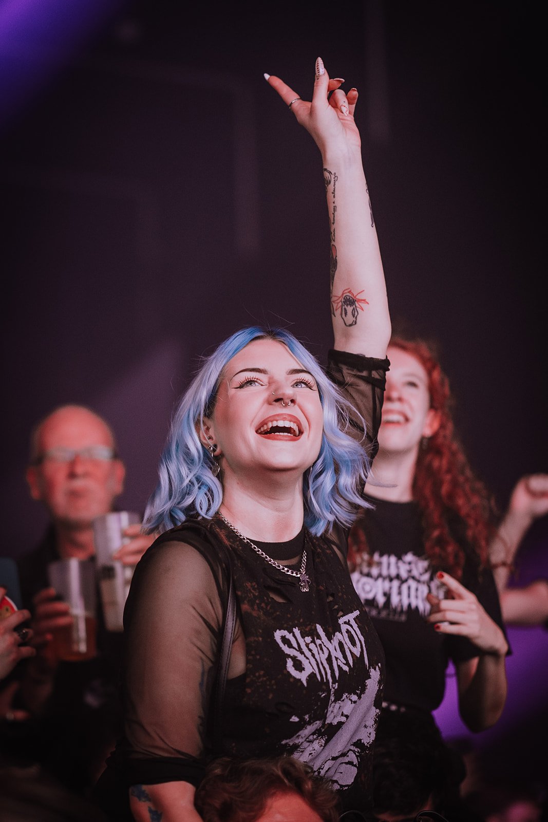 A woman with blue hair, smiling and raising her hand at a concert or event. She is wearing a black Slipknot t-shirt and has tattoos on her arm. Other people are visible in the background.