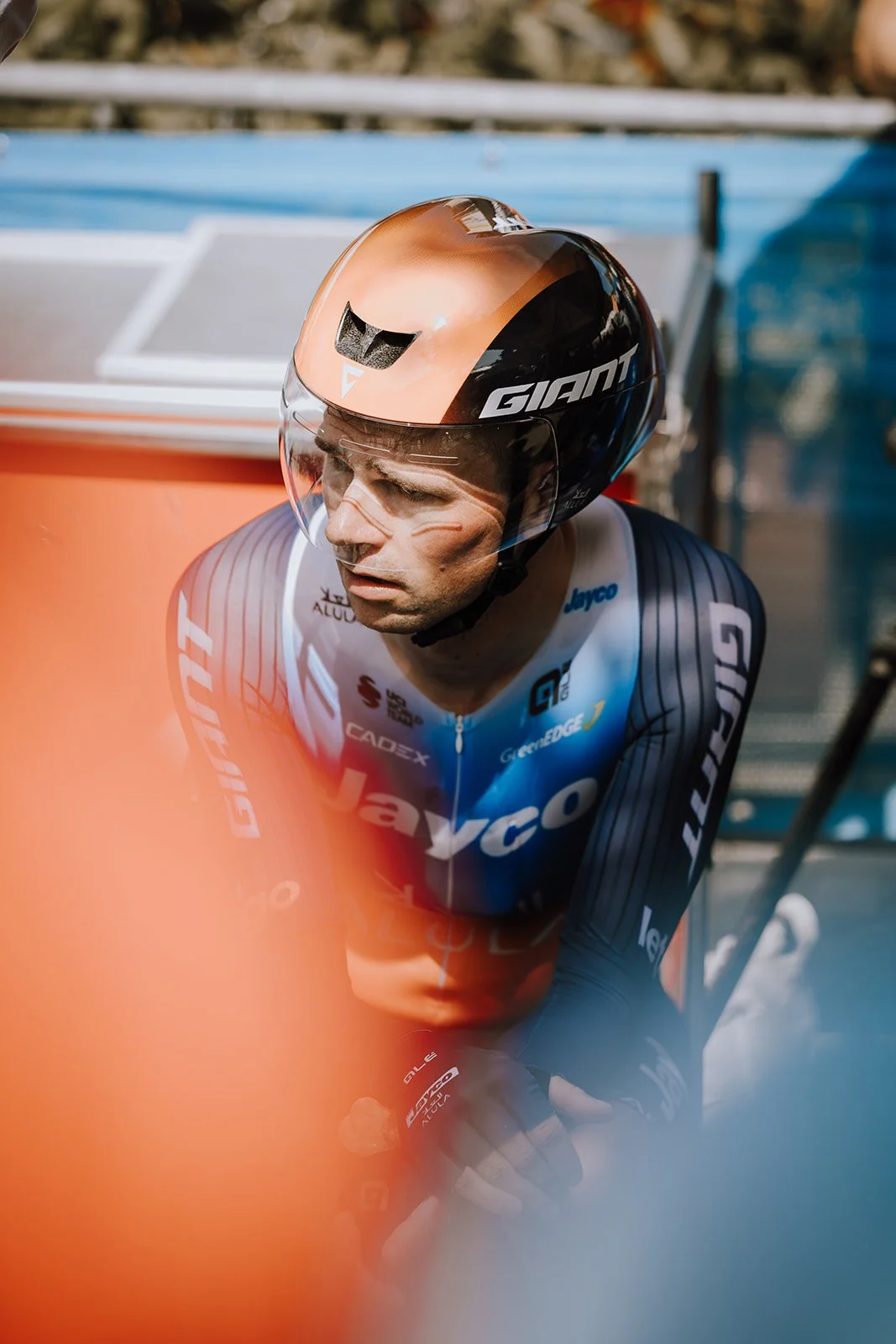 A male cyclist wearing a blue and black Jayco racing suit, black gloves, and a black and copper helmet with the brand name GIANT, sitting on a bench or stairs, looking downward.
