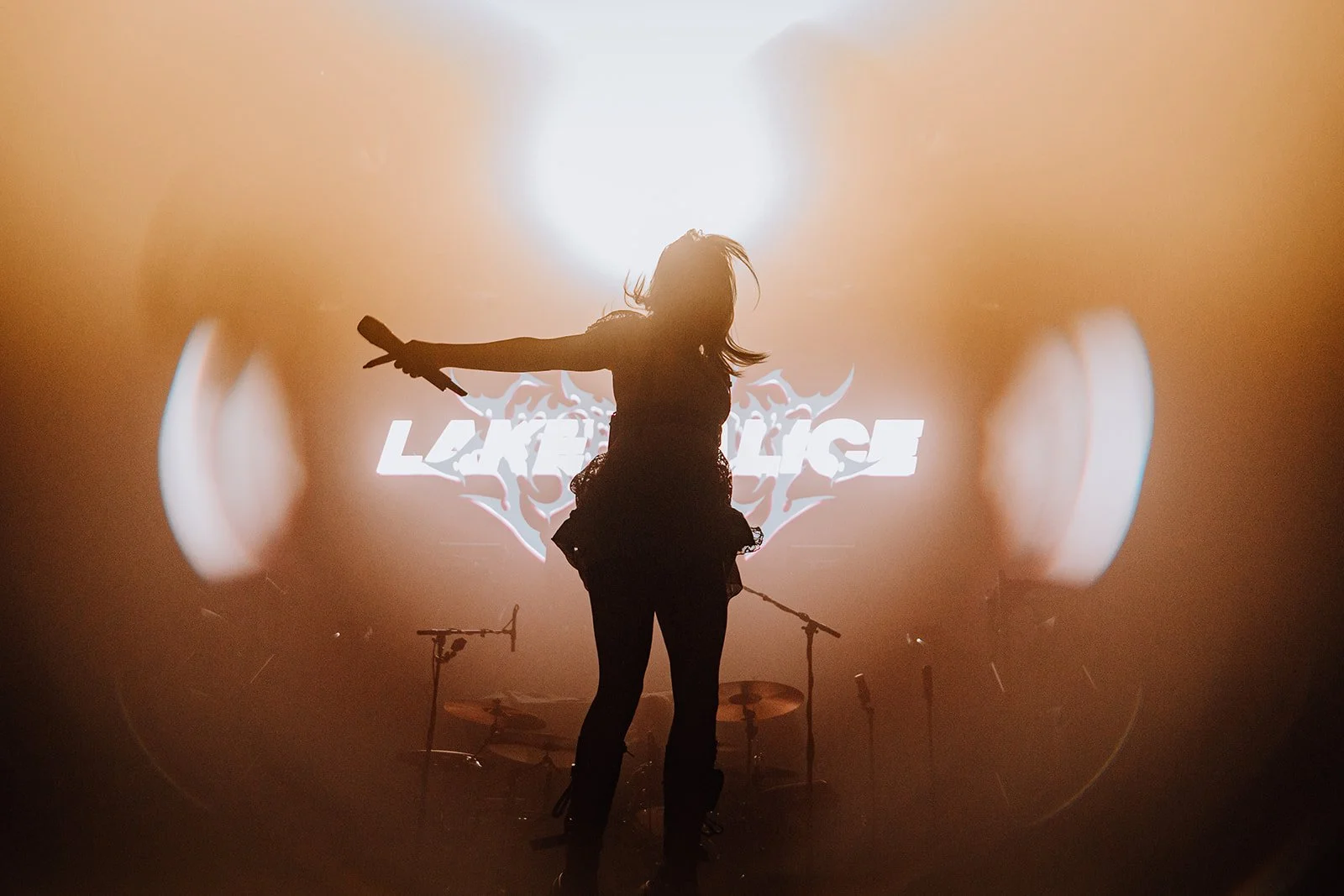 Silhouette of a female singer performing on stage with arms outstretched, backlit by bright stage lights, with a band and a large screen displaying the word "Lament" in the background.