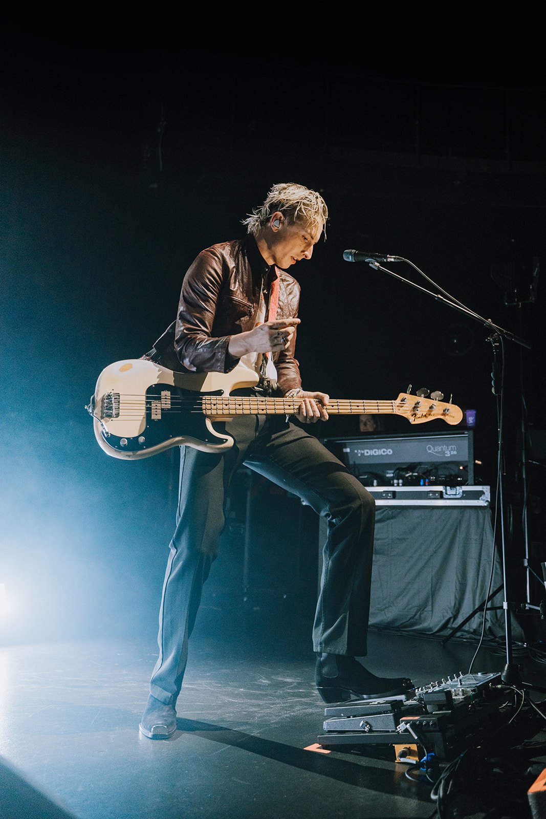 A musician playing an electric bass guitar on stage, wearing a brown leather jacket and black pants, with pedal effects and audio equipment in the background.