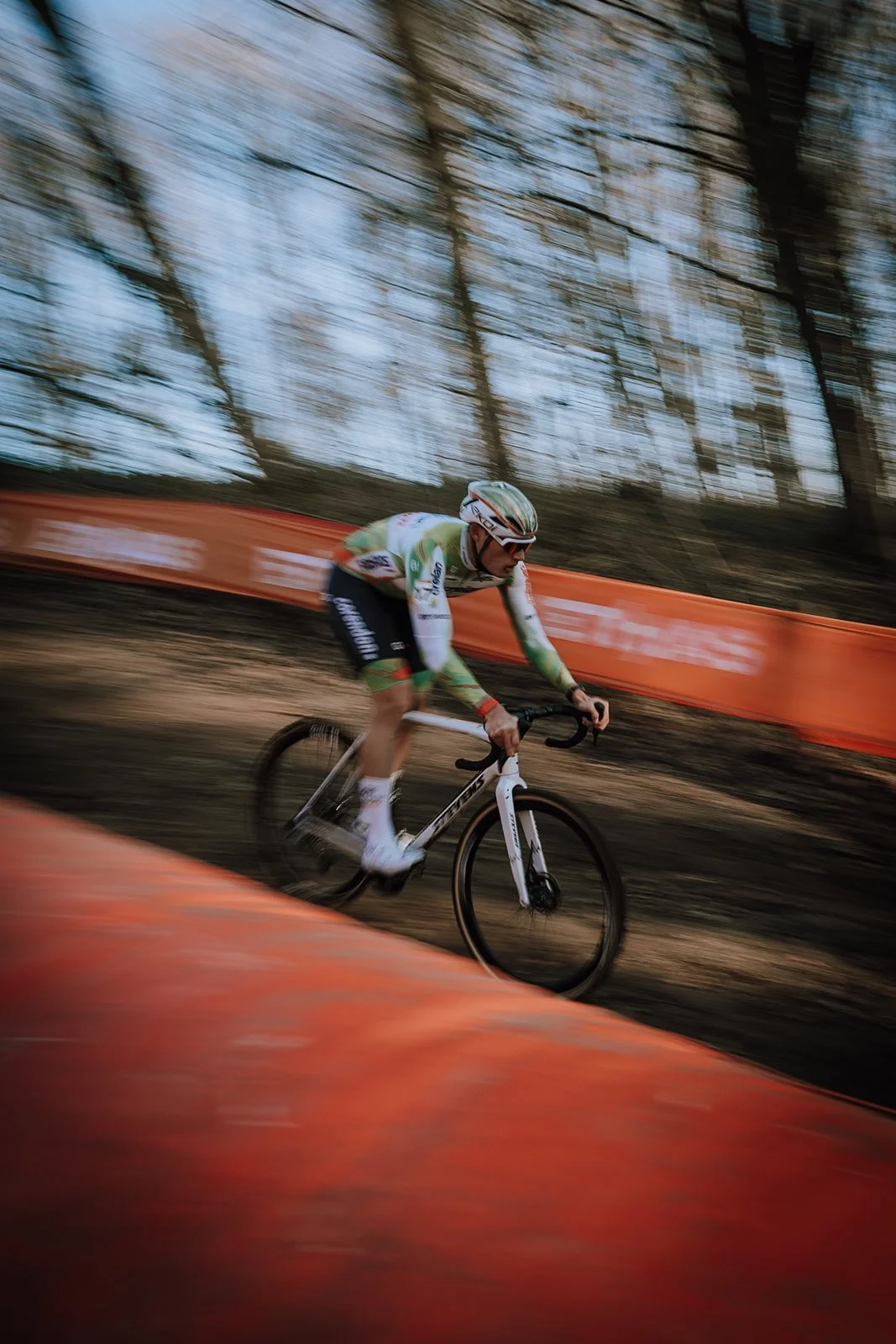 Cyclist riding a bike on a dirt trail during a race in a wooded area with orange safety barriers along the path.