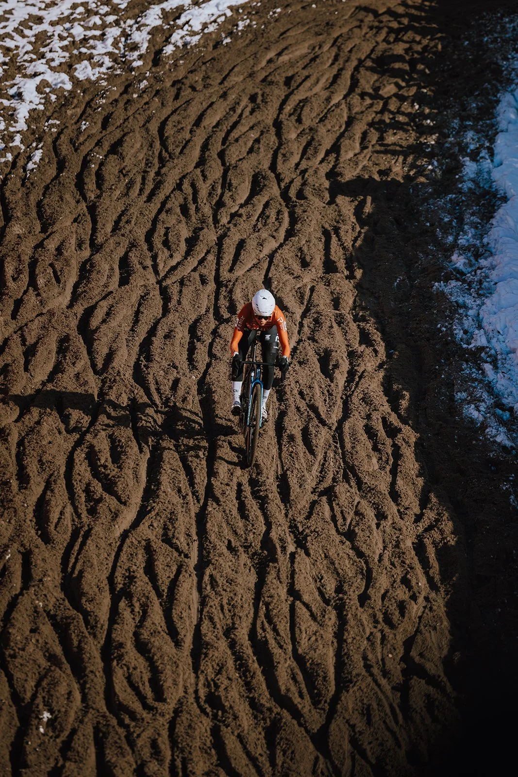 A cyclist riding a mountain bike on a dirt trail with tire tracks and patches of snow on the sides.