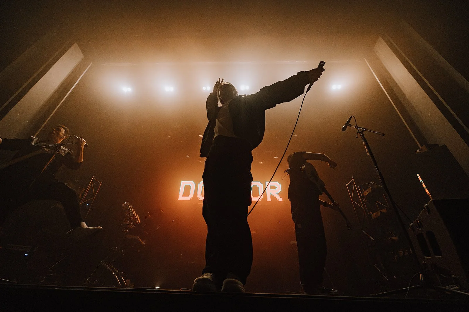 Silhouettes of people on stage during a concert, one person holding a microphone, another with a handheld microphone, with colored stage lighting and a large screen displaying 'DOR' in the background.
