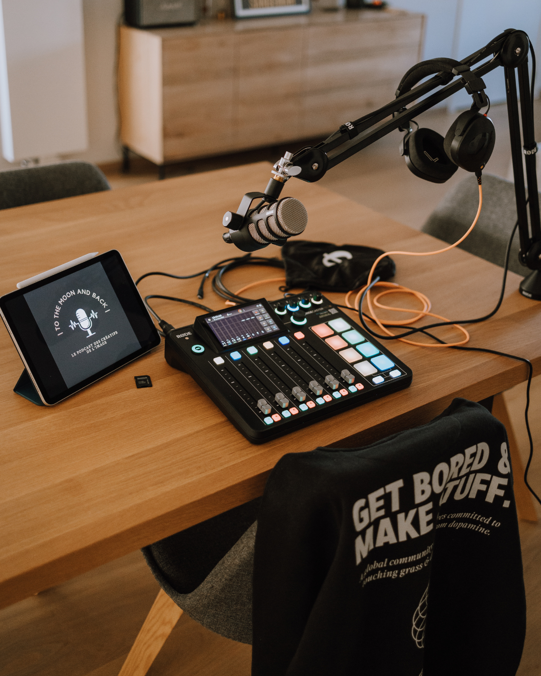 A podcast recording setup on a wooden table with a tablet displaying the podcast logo, a digital audio mixer, a microphone mounted on an adjustable arm, and headphones placed nearby.
