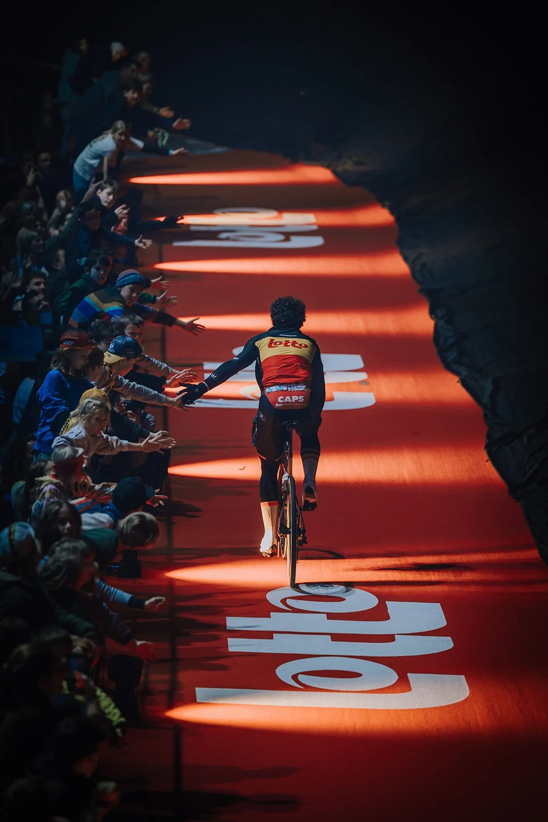 A cyclist riding through a tunnel with an enthusiastic crowd reaching out to high-five on the sides. The tunnel is illuminated with red lighting and the floor displays white and red advertising text.