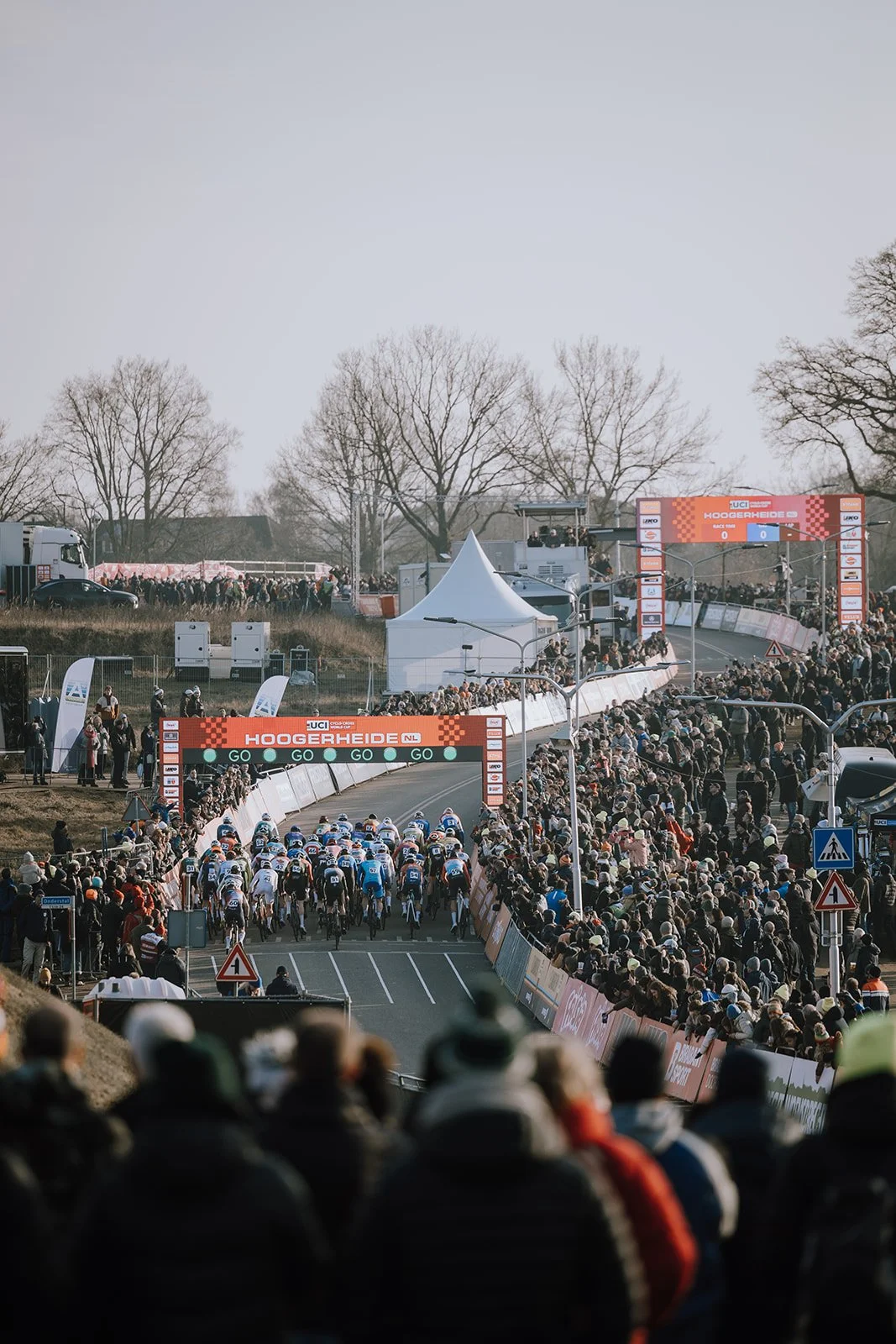 Cyclists racing on a closed road with crowd on both sides, starting a race at Hoogereide, with banners and a large digital scoreboard overhead.
