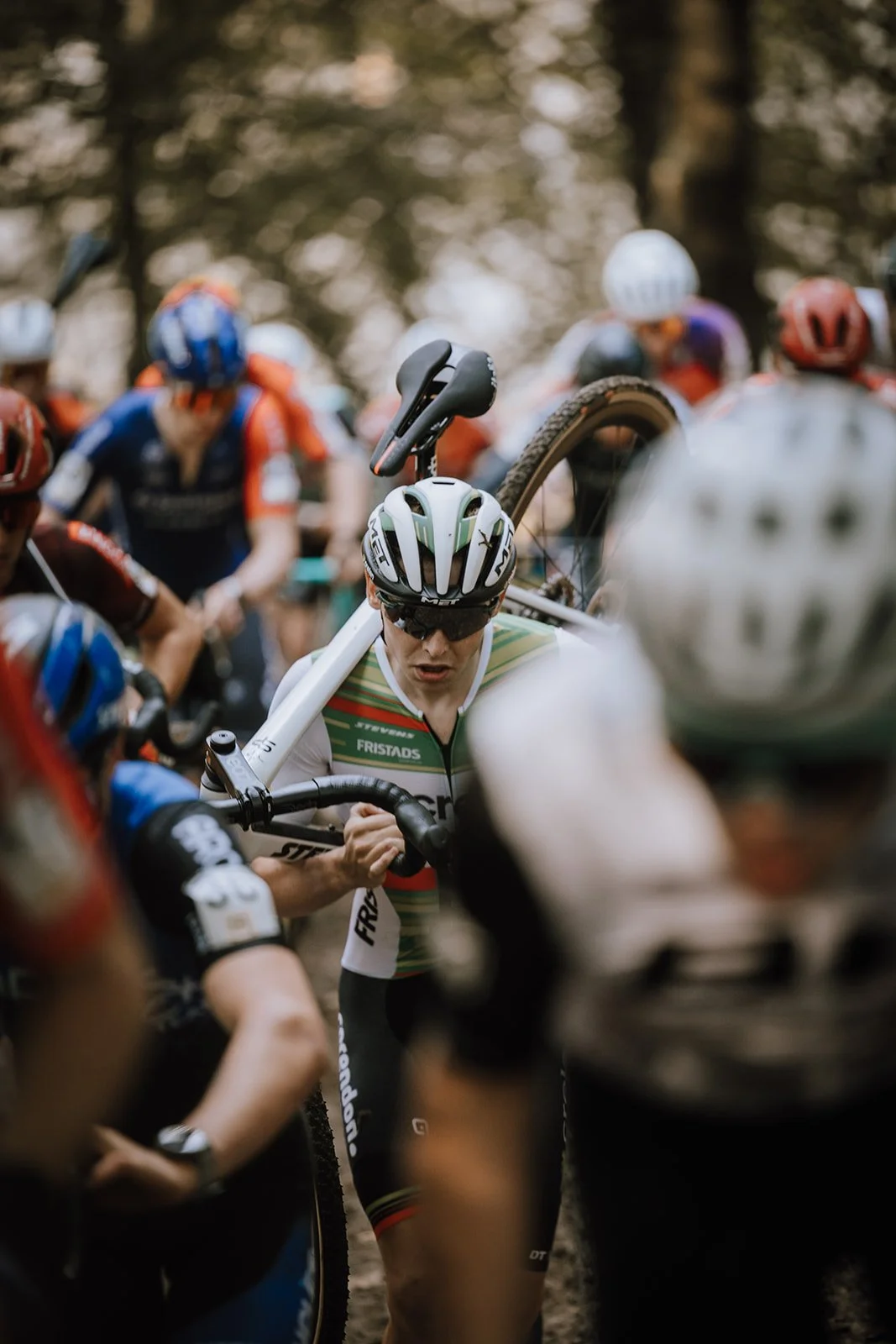 A cyclist in a white and black jersey and helmet carrying his bike over his shoulder through a crowded outdoor event.