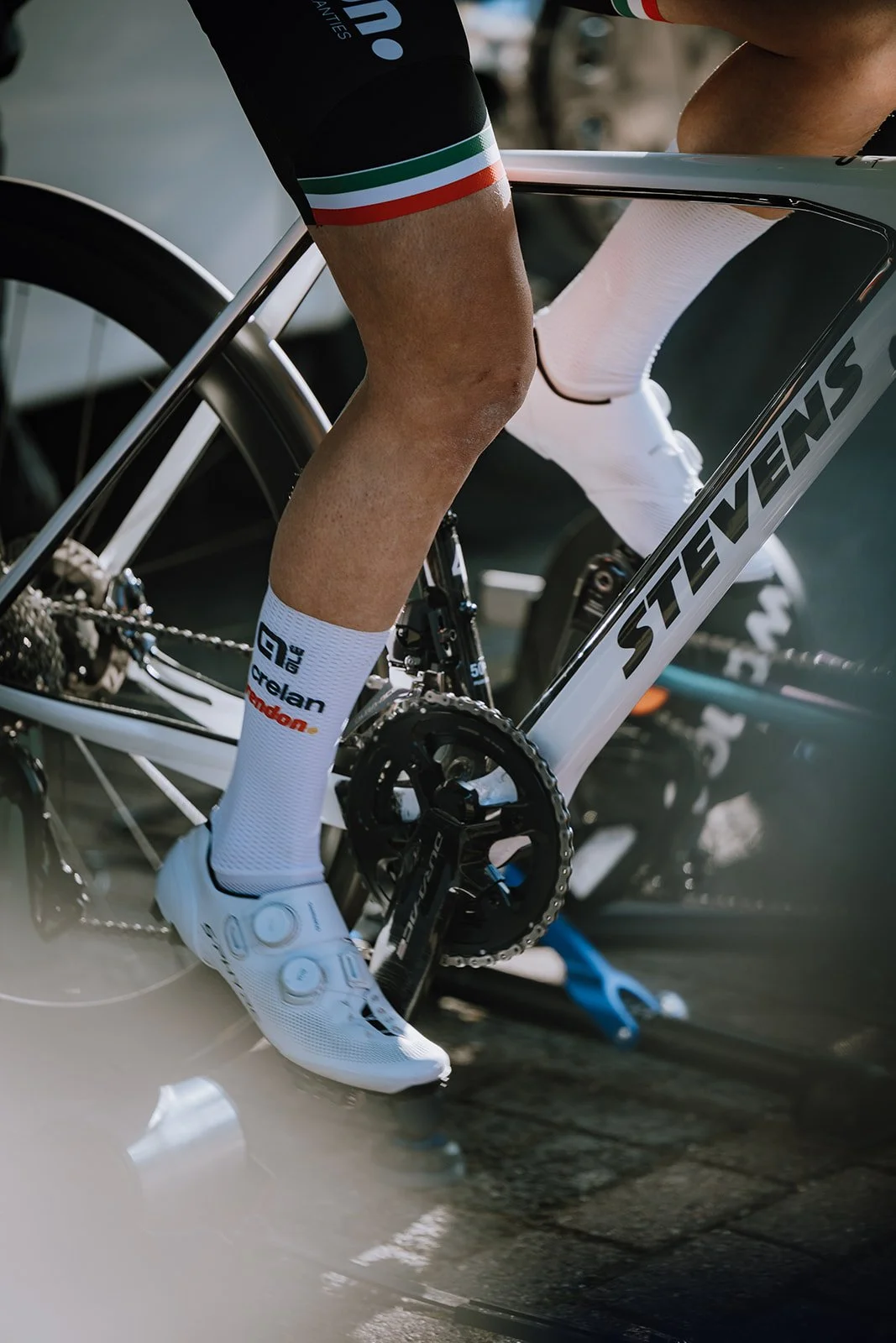 Close-up of a cyclist's legs on a STEVENS racing bicycle, wearing white cycling shoes and socks, near the pedals and chainring.
