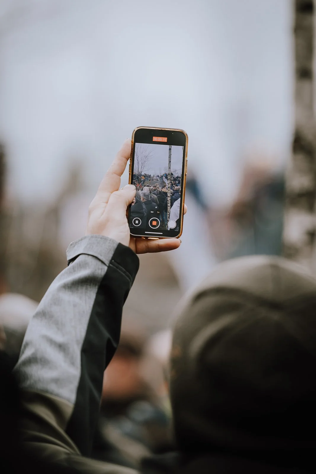 Person holding a smartphone recording a crowd at an outdoor event on a cloudy day.