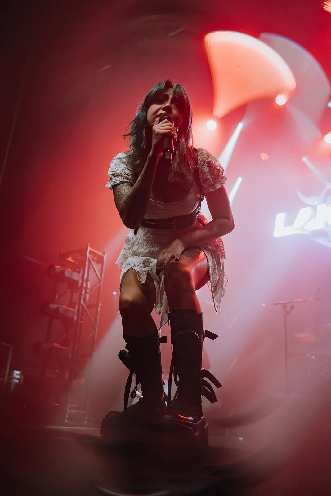 A female singer performing on stage during a concert, illuminated by red and white stage lights, wearing a lace dress and black knee-high boots, with tattoos visible on her arm.