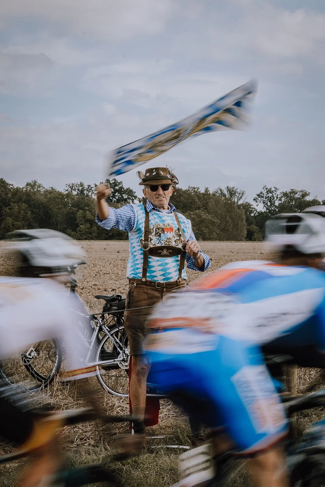 A man dressed for Oktoberfest, wearing traditional Bavarian clothing including lederhosen, standing in a field with bicycles around him, waving a Bavarian flag.