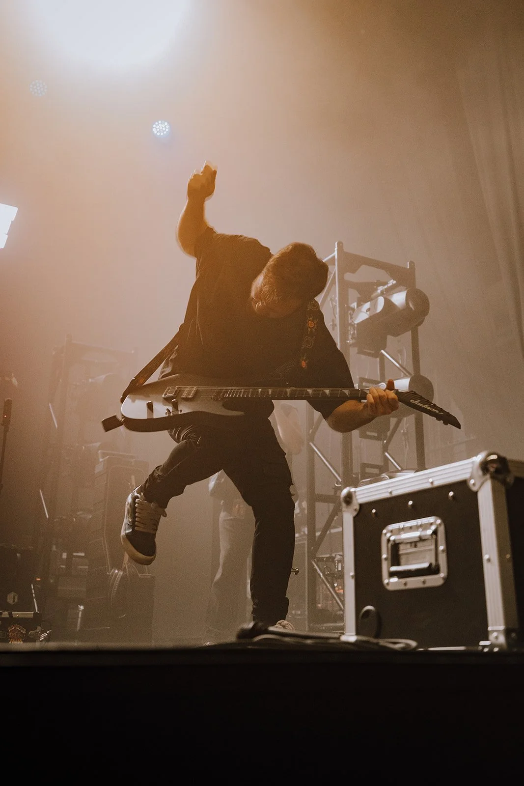Guitarist performing on stage with stage lights and equipment visible in the background.