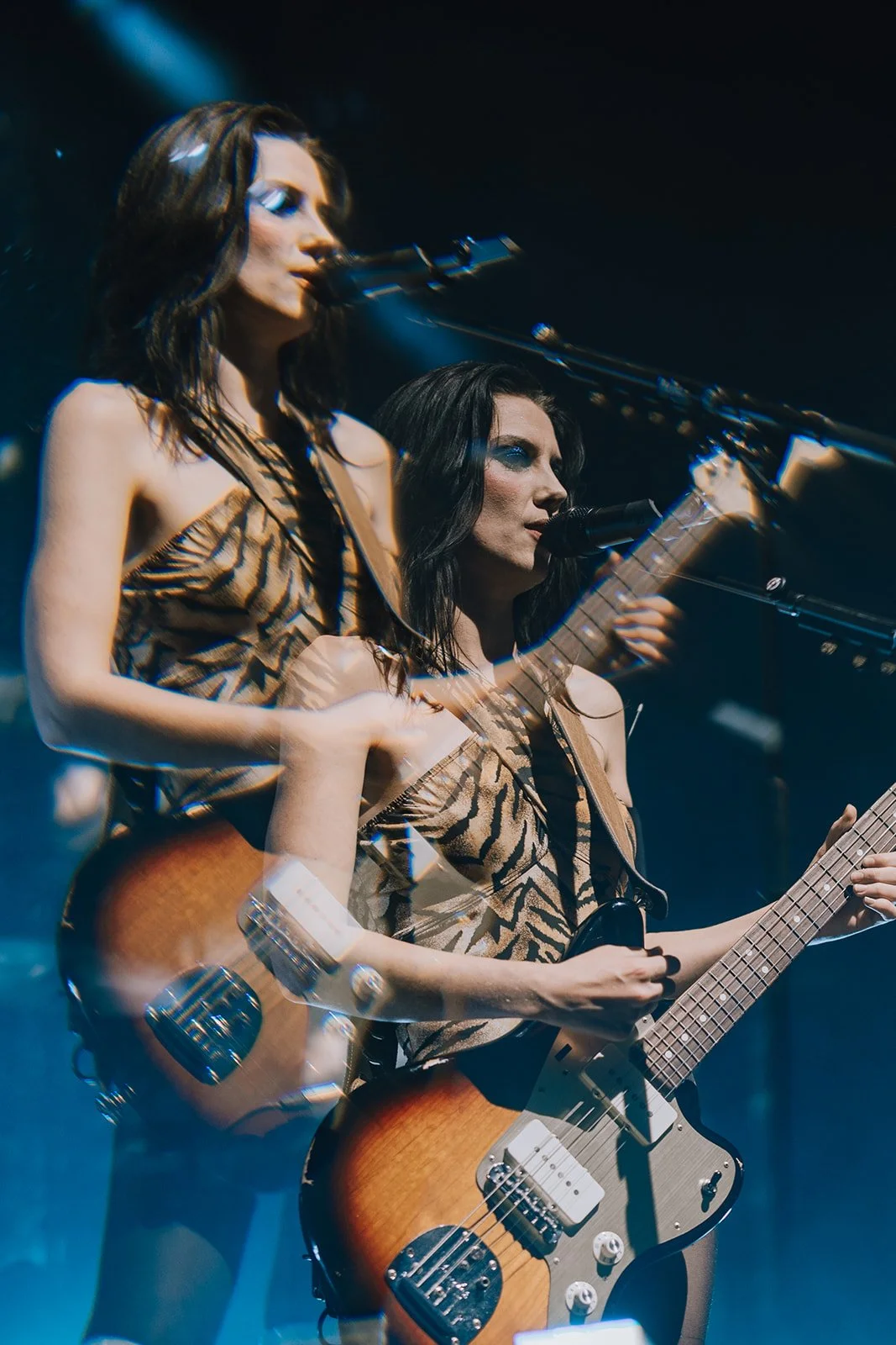 Two female musicians performing on stage with guitars, one singing into a microphone and the other singing into a microphone while playing guitar, both wearing animal print tops.