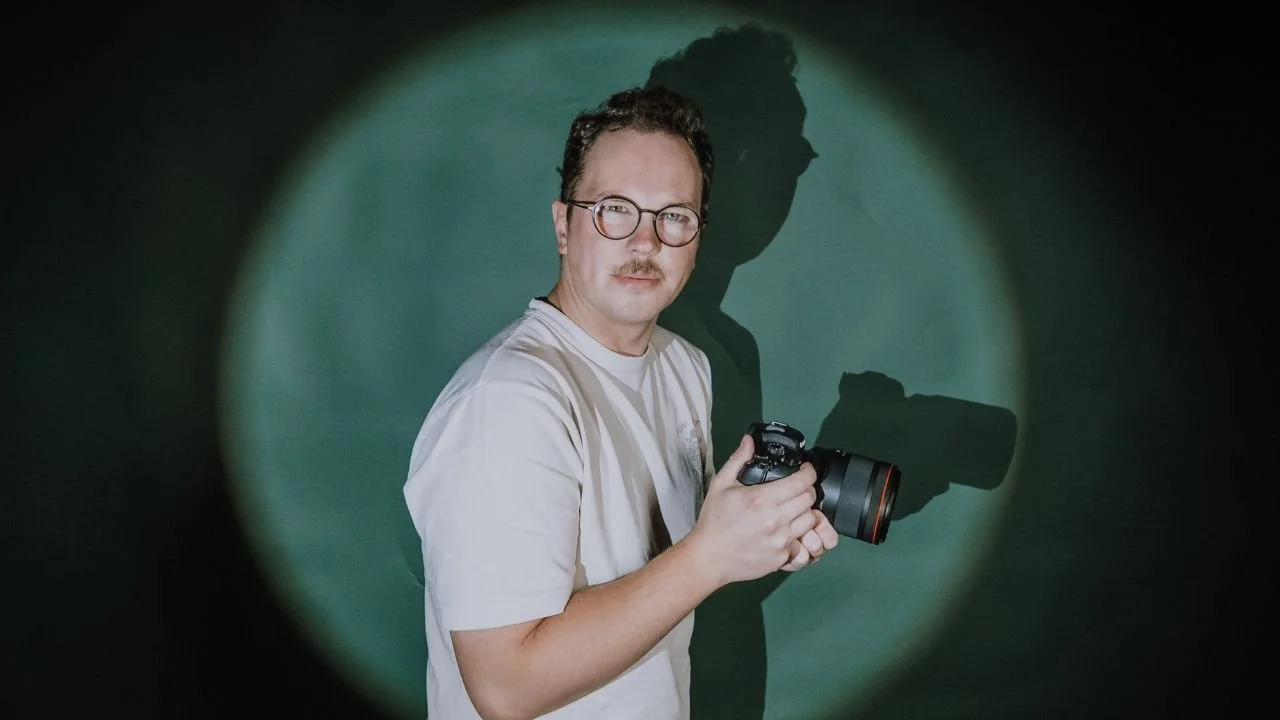 A man with glasses and a mustache holding a camera, standing in front of a green screen with a spotlight creating a shadow.