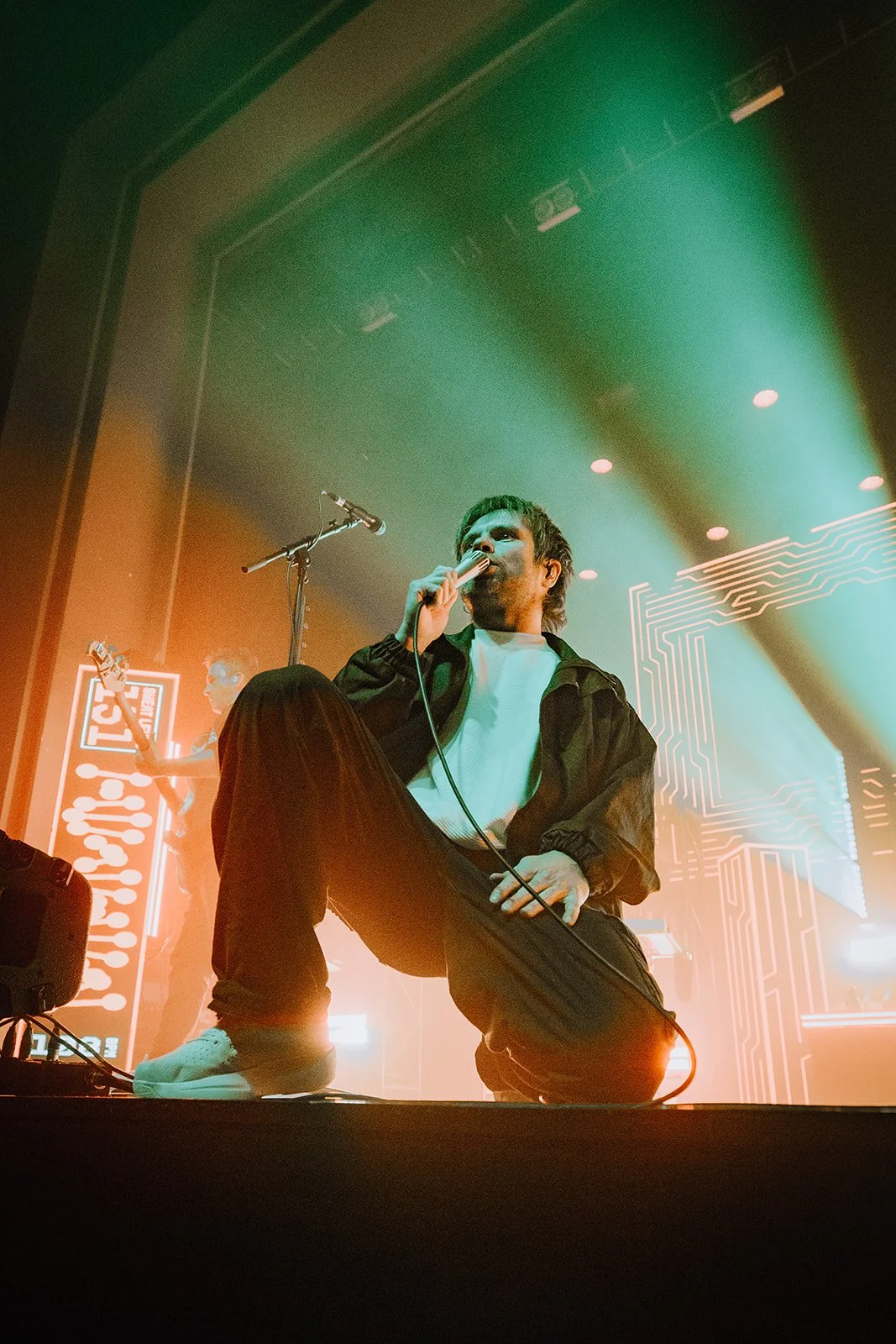 Kneeling male singer holding a microphone on stage with colorful lighting and digital graphics in the background.